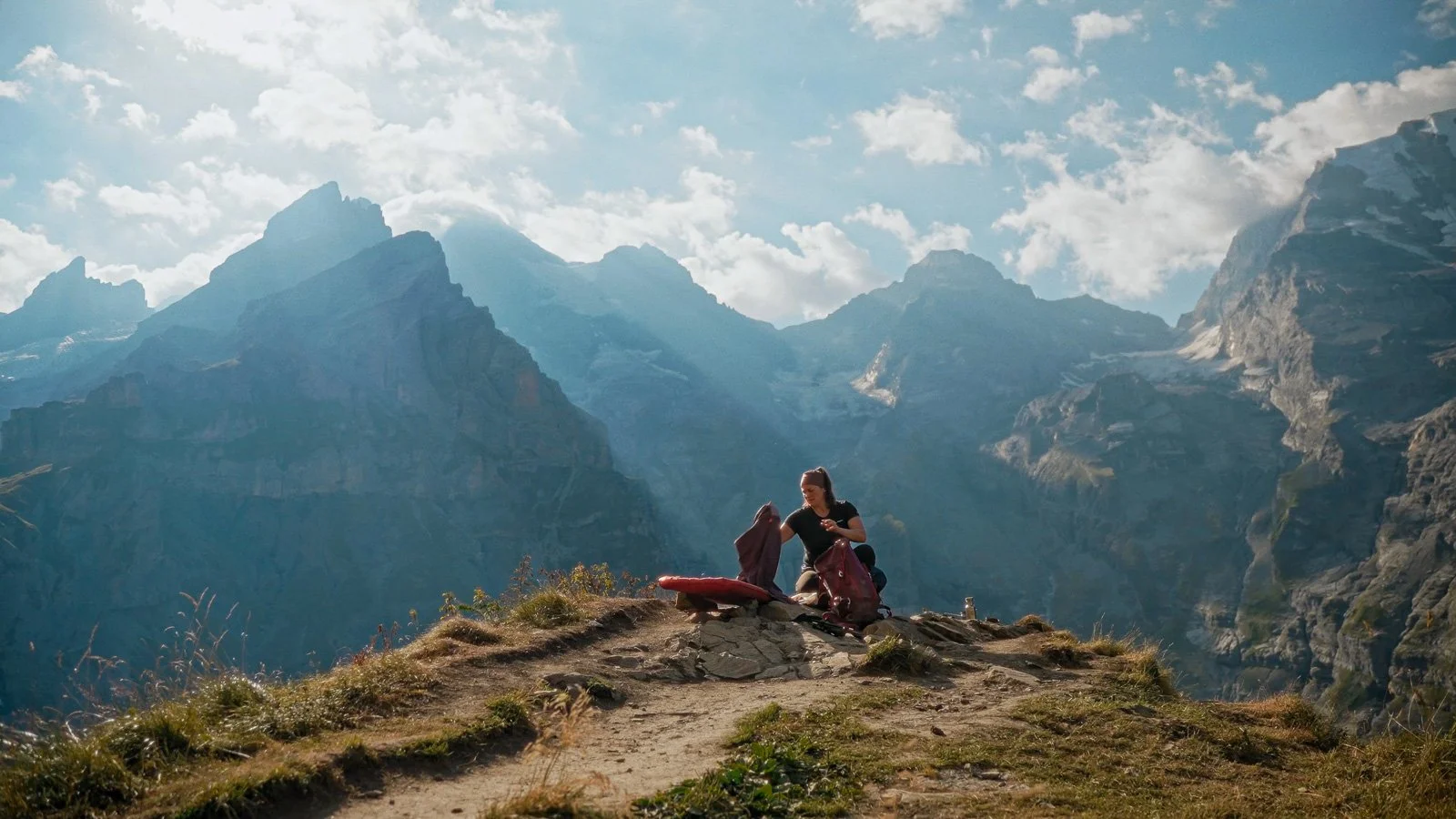 A woman sitting on a small mound of dirt and grass in a mountainous landscape, packing or unpacking gear with towering mountain peaks in the background and a partly cloudy sky.