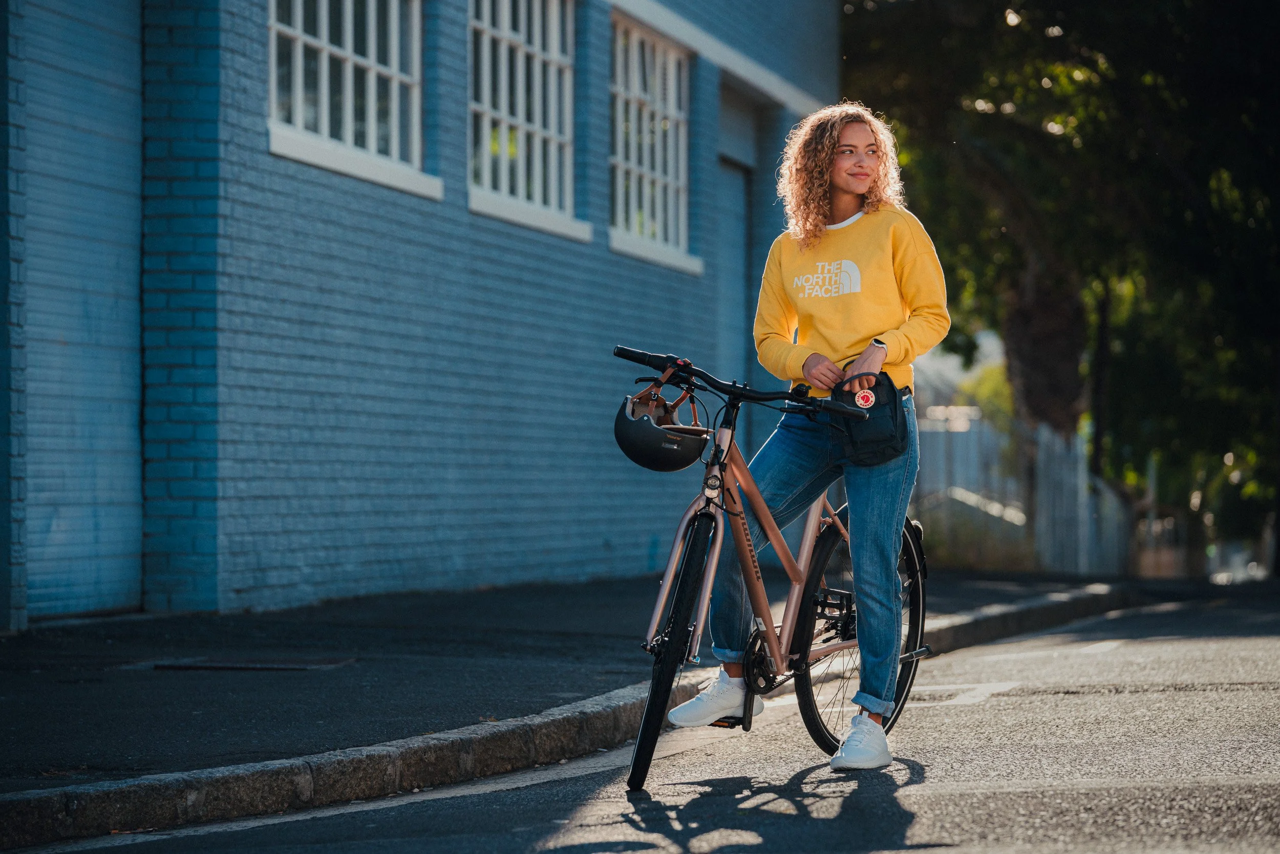 A young woman standing next to her bicycle on a city street, wearing a yellow North Face sweatshirt, blue jeans, and white sneakers, smiling as the sun sets in the background.