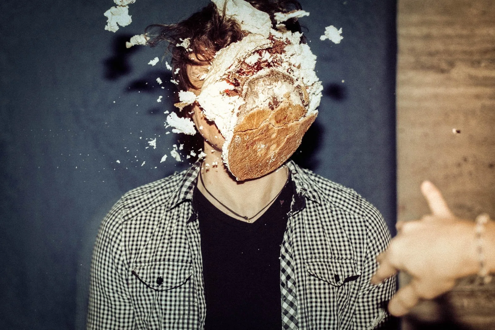 A person with a checkered shirt has a loaf of bread covering their face, smashing bread into their face, with crumbs and pieces flying in the air.