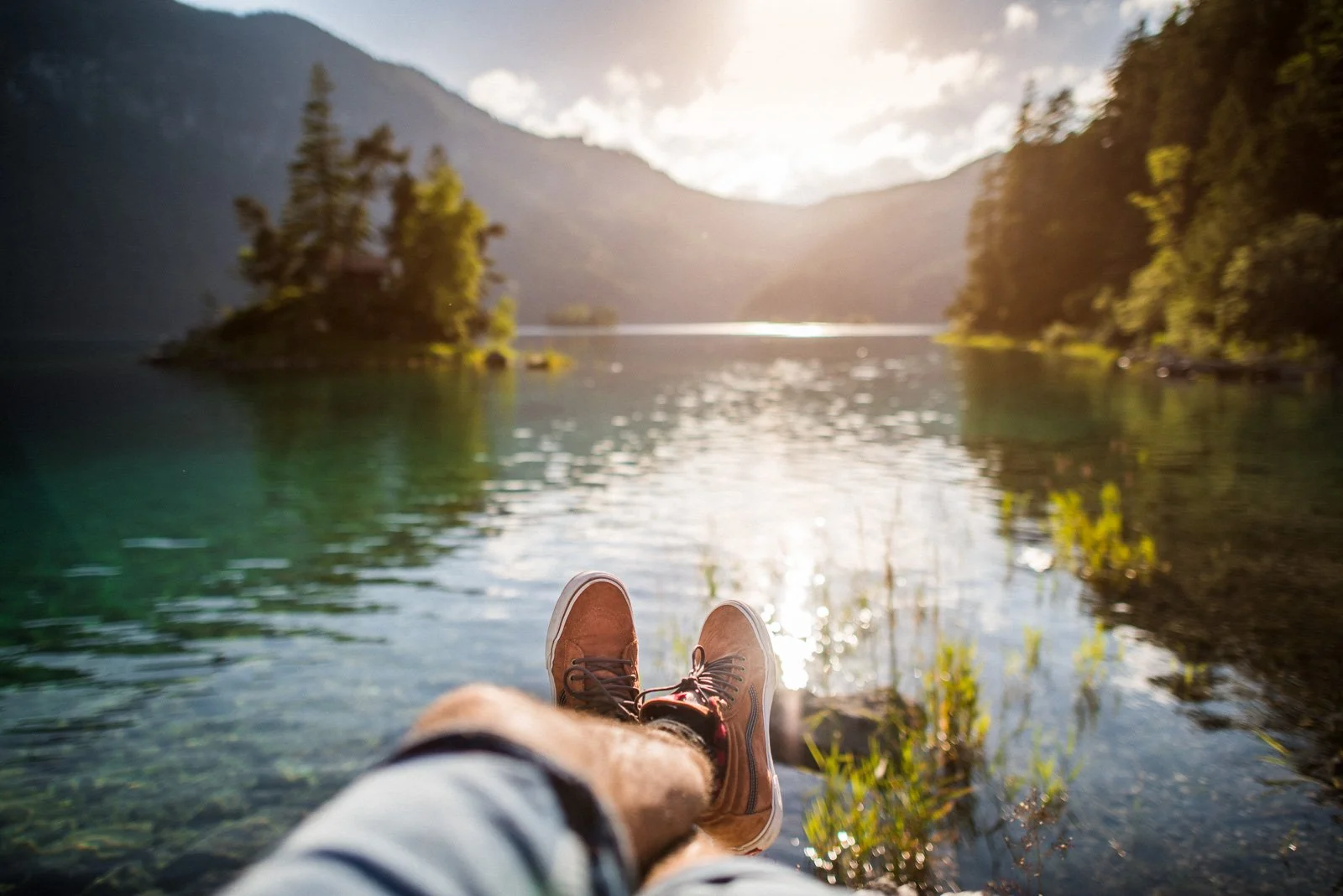 Person with brown shoes and shorts sitting on a lakeside, overlooking a tranquil body of water with trees and mountains in the background during sunset.