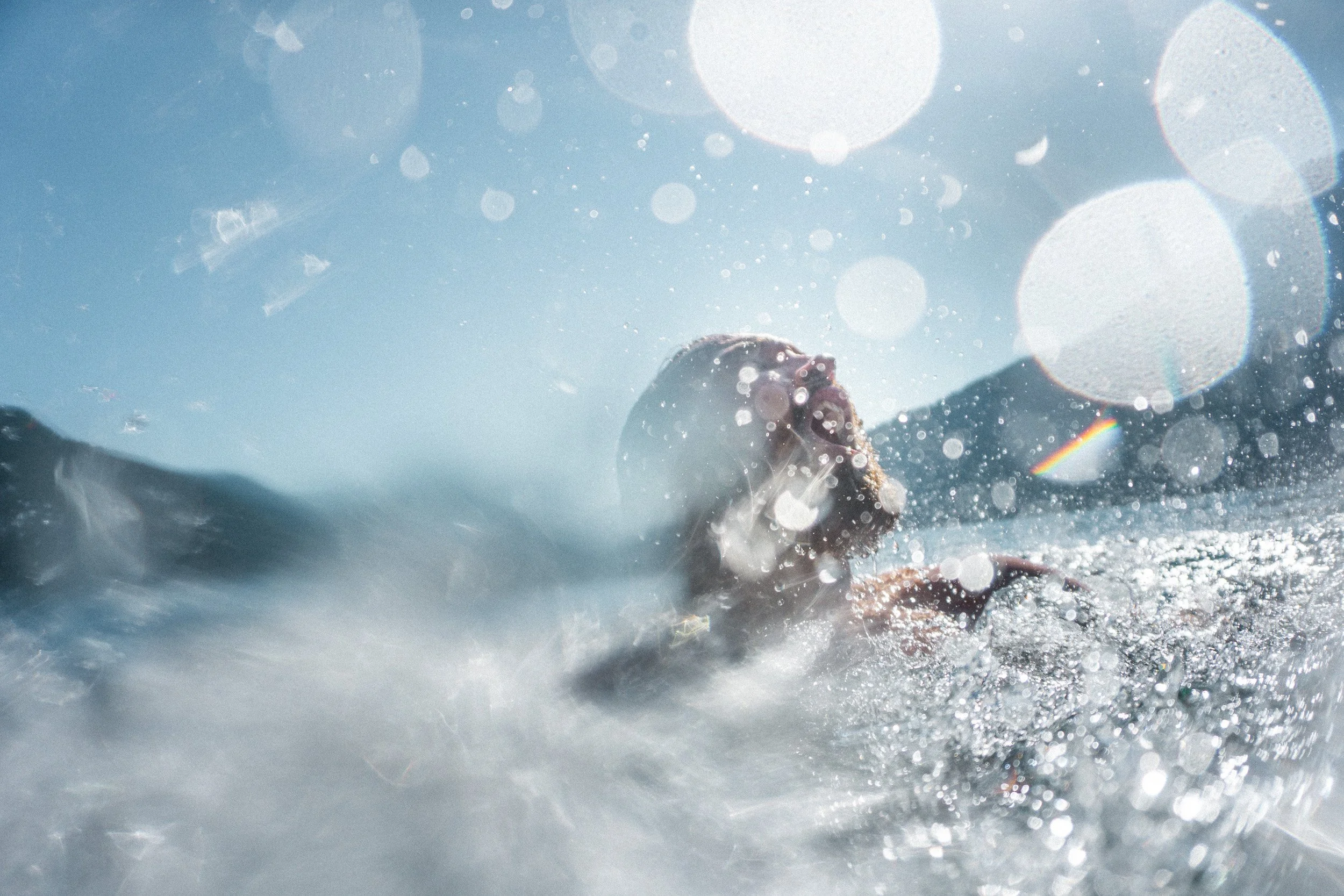 Person swimming in the ocean with water splashing around and sunlight creating lens flare effects.