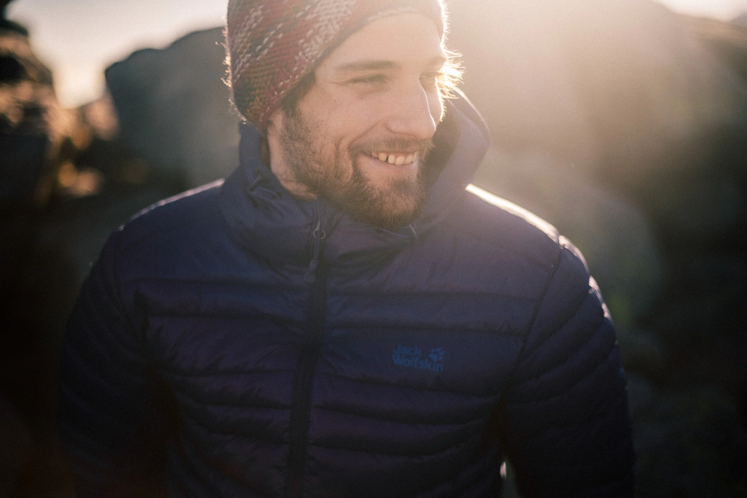 A man wearing a dark blue Jack Wolfskin jacket and a colorful knitted hat smiling outdoors in sunlight.