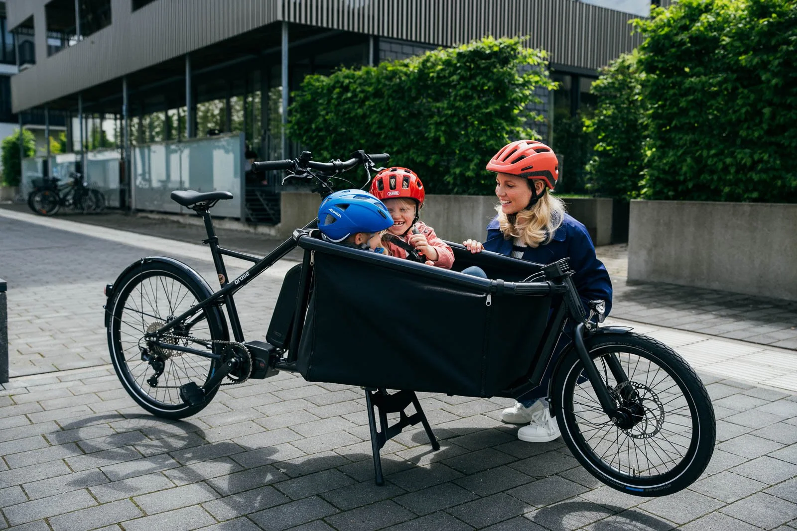 Three people, two children and one adult, wearing helmets and smiling around a black cargo bike with a large compartment in front, on a city sidewalk with trees and modern buildings in the background.
