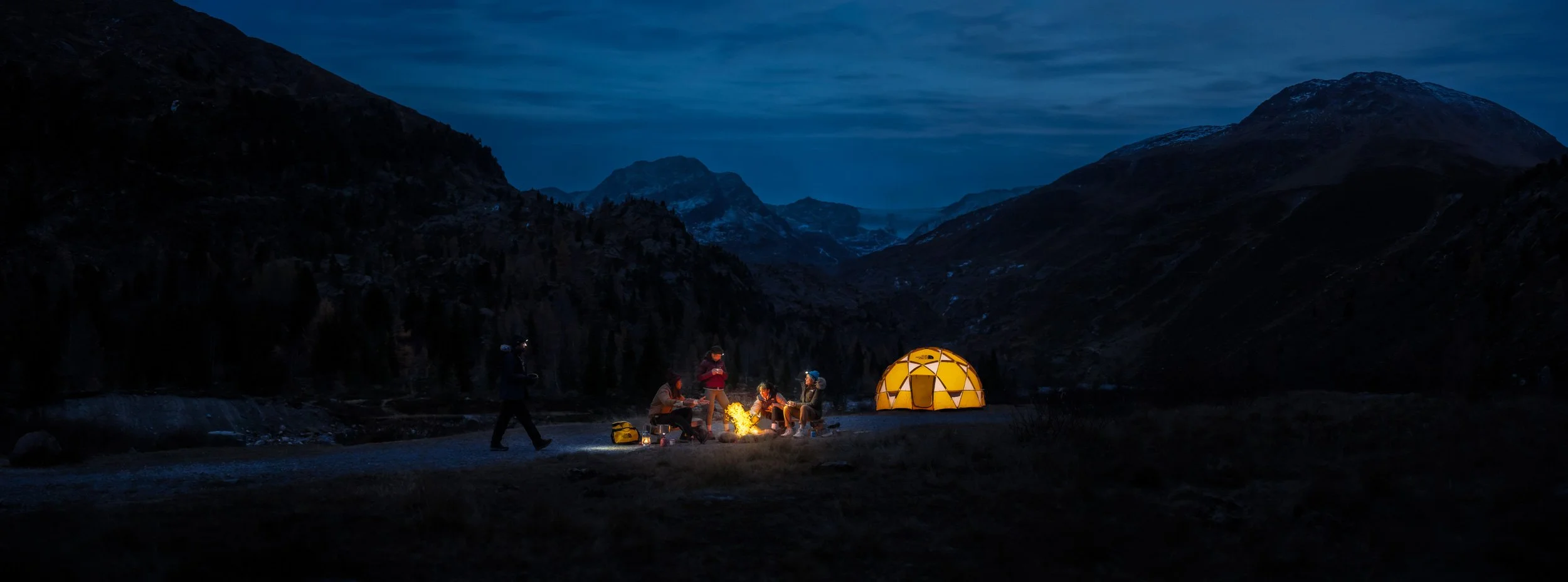 A nighttime mountain camping scene with a group of people around a campfire, a lit yellow tent, and a dark mountain landscape in the background.