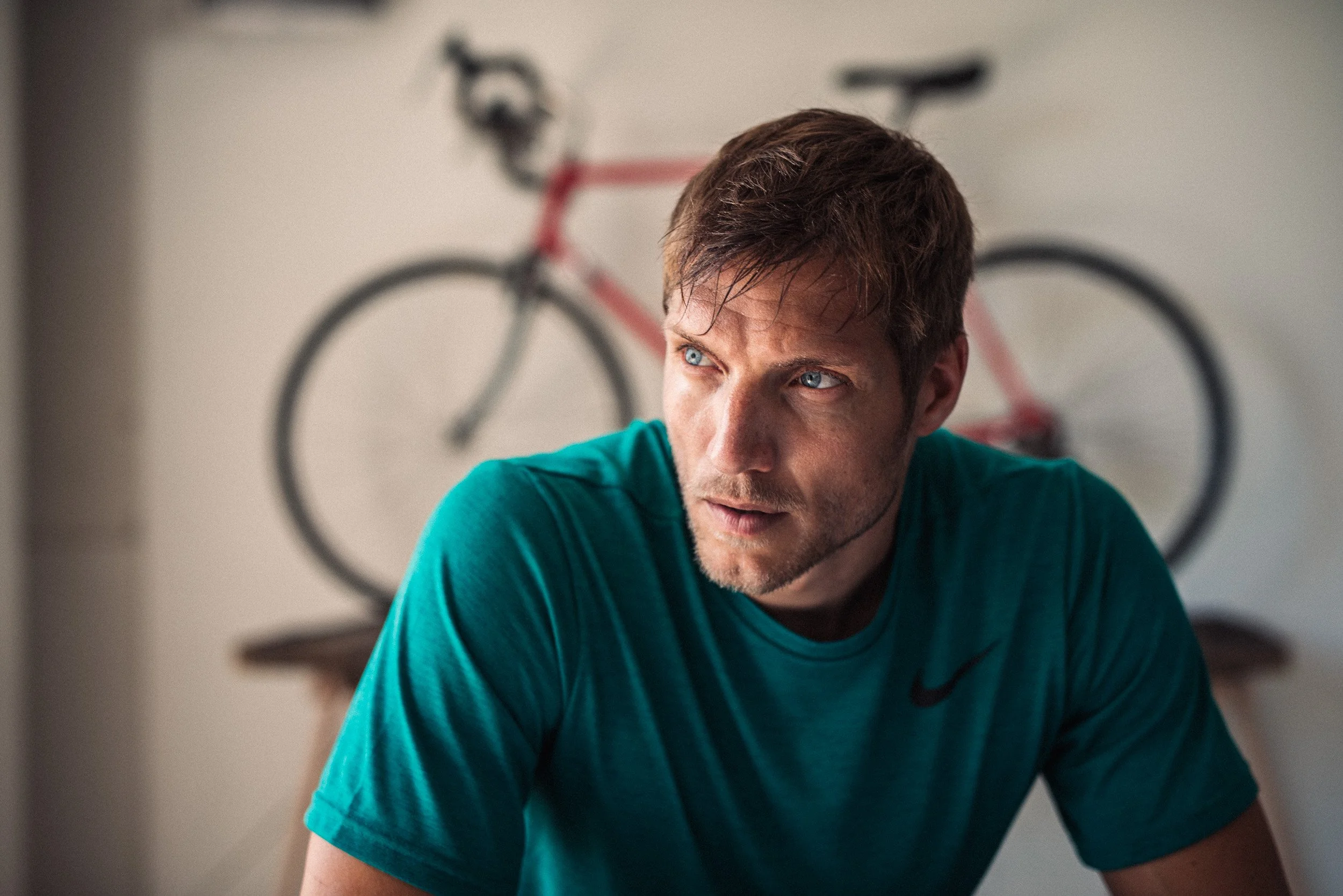 A man in a teal Nike shirt with brown hair and blue eyes looking to the side with a bicycle on the wall behind him.