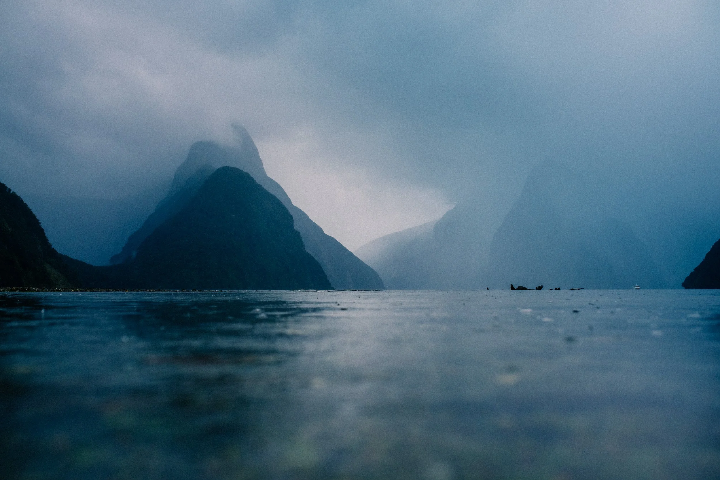 A cloudy, misty scene of a large body of water with tall, steep mountain cliffs on both sides, partially obscured by fog.