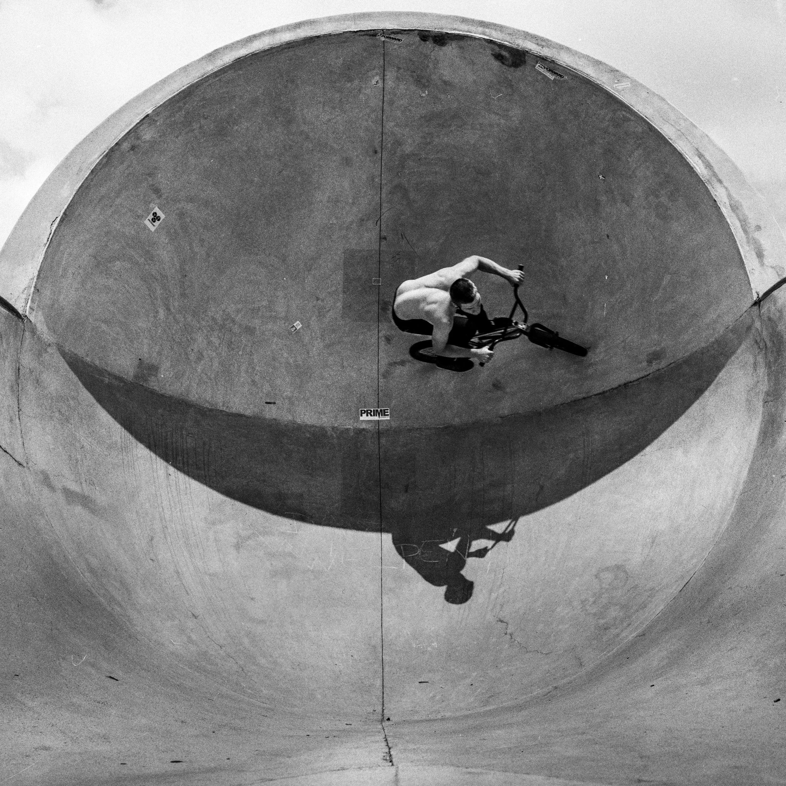 A shirtless man riding a BMX bike inside a concrete skate park bowl, with his shadow visible on the surface.