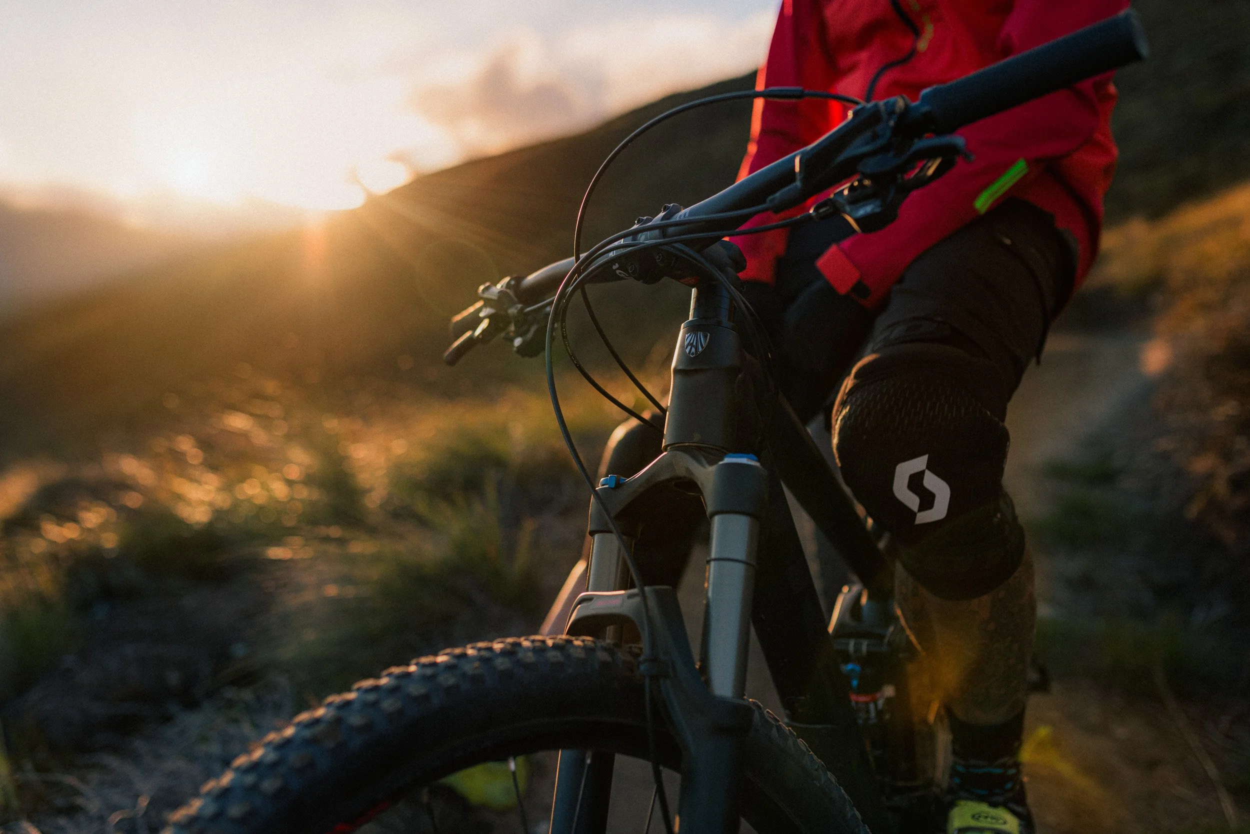 Close-up of a mountain biker in a red jacket riding on a trail during sunset, with the sun low over a hilly landscape.