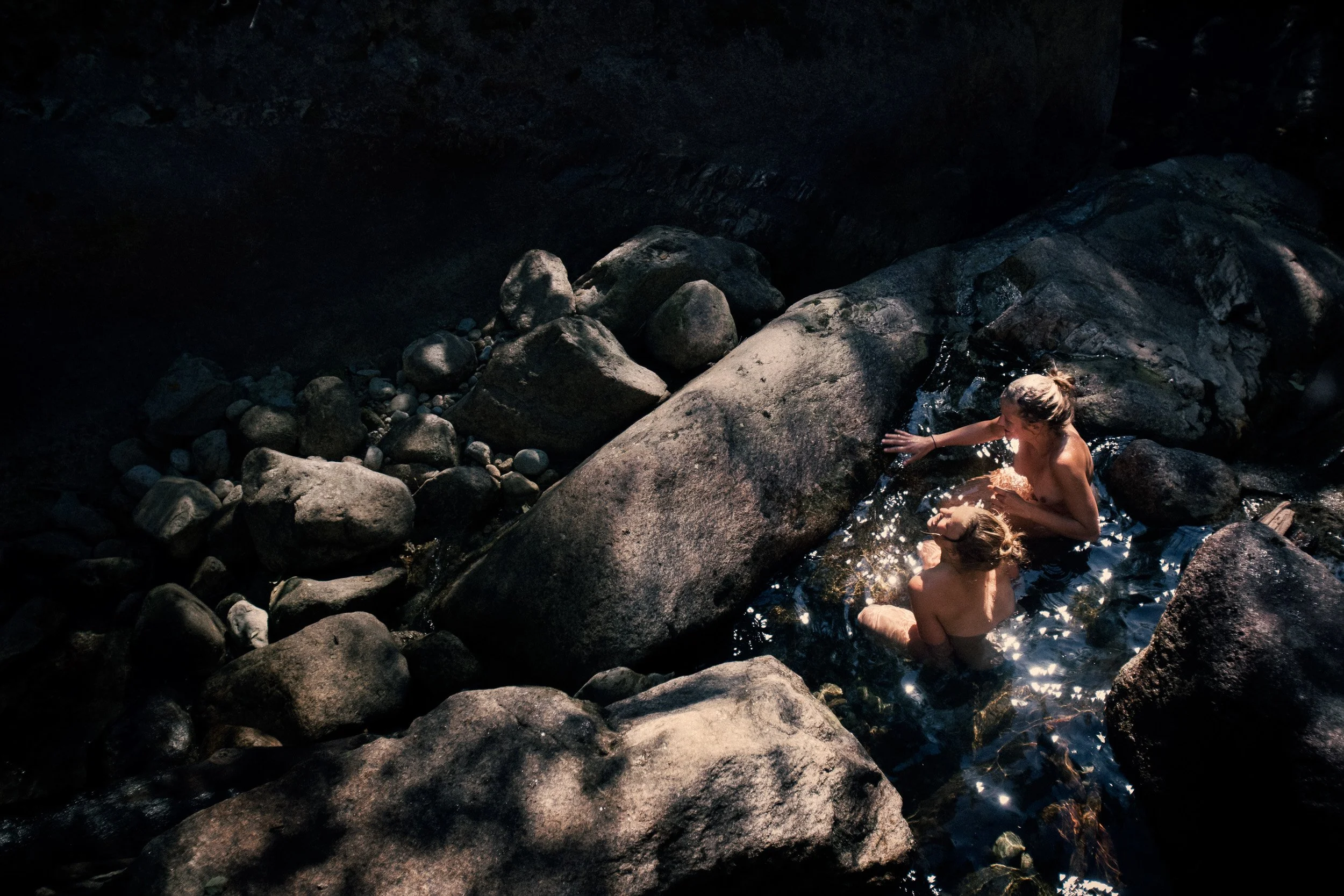 Two people, a woman and a child, are sitting in a small, natural water pool surrounded by large rocks.