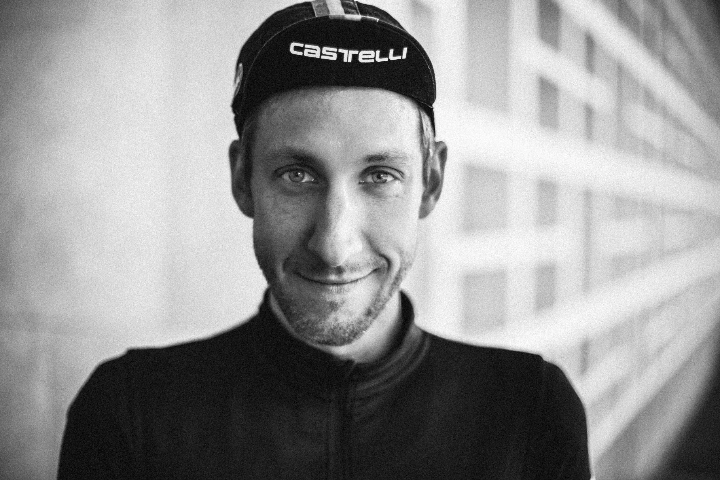 Black-and-white photo of a smiling man wearing a Castelli cap and a cycling jersey, standing in front of a brick wall.