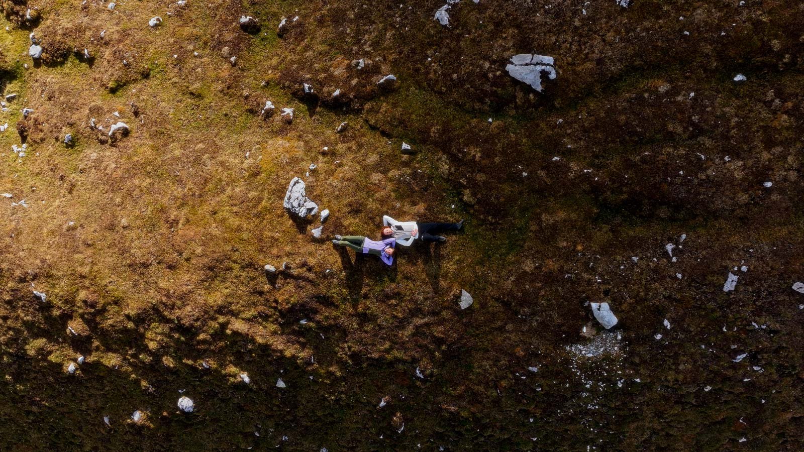 Aerial view of two people lying on a mossy rocky hillside, facing each other.