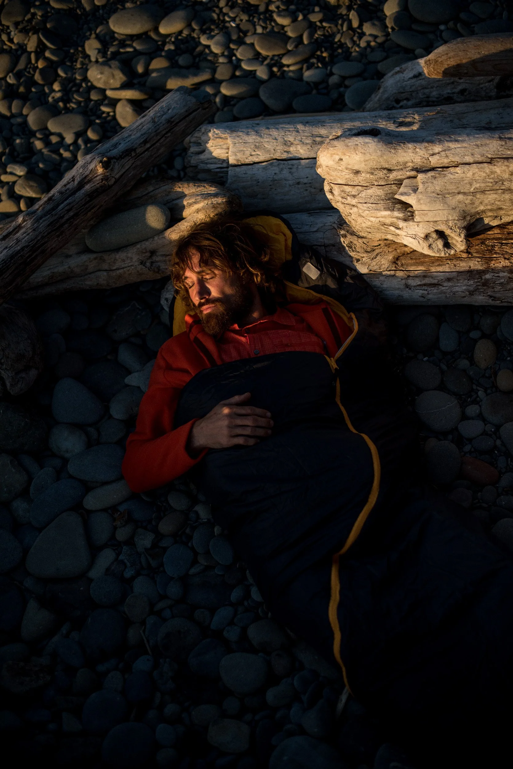 A man sleeping on a rocky beach surrounded by driftwood, with his hand resting on his chest, wearing a red jacket and black sleeping bag.