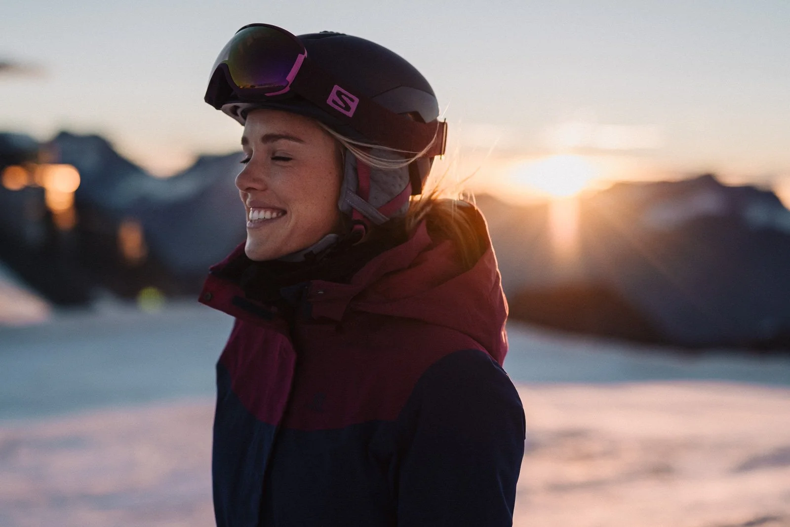 A woman wearing a ski helmet and goggles smiles with her eyes closed during sunset in a snowy mountain landscape.