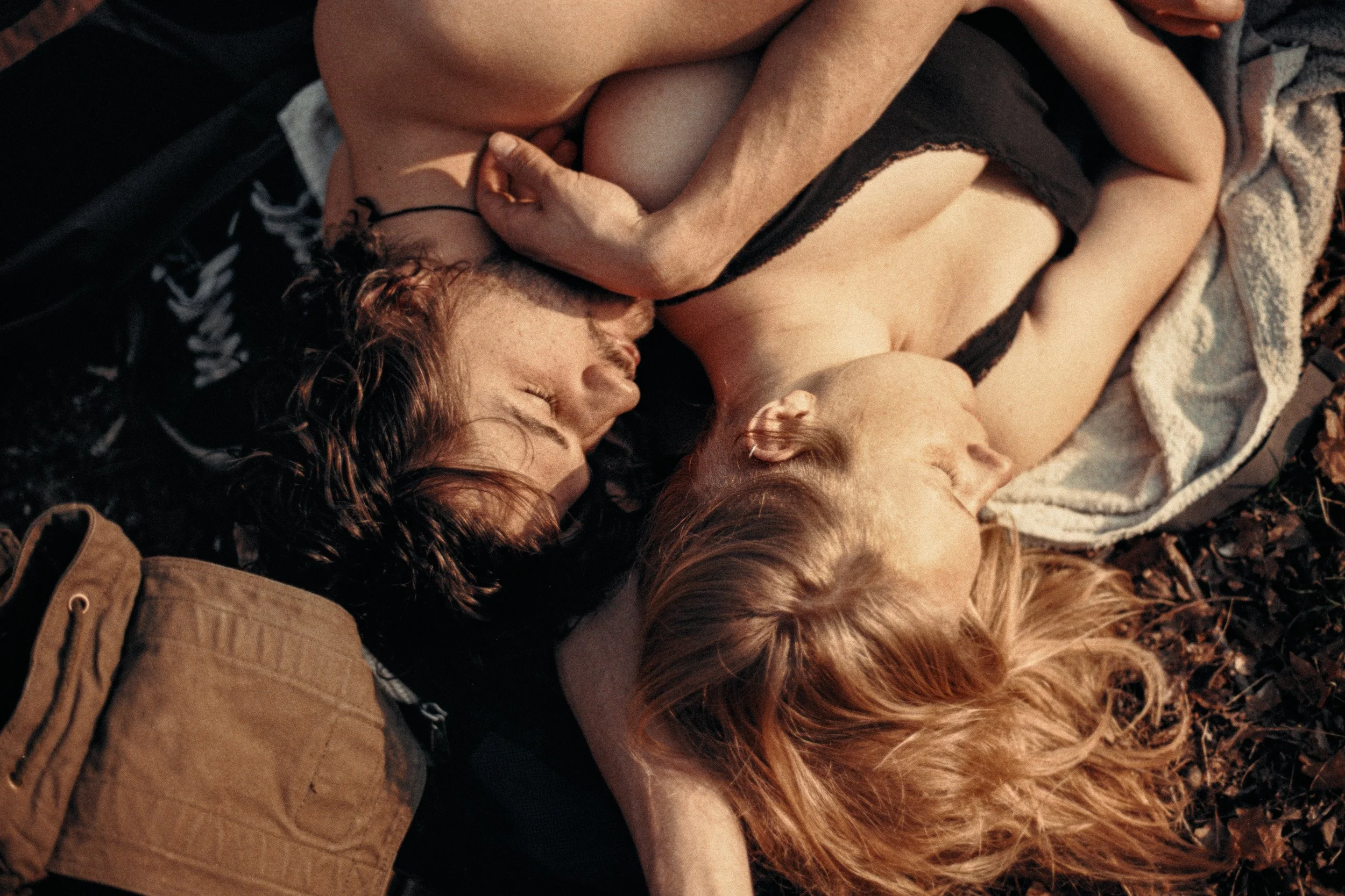 Two women lay close together outdoors on a blanket, embracing each other with their eyes closed, in a warm and intimate moment.