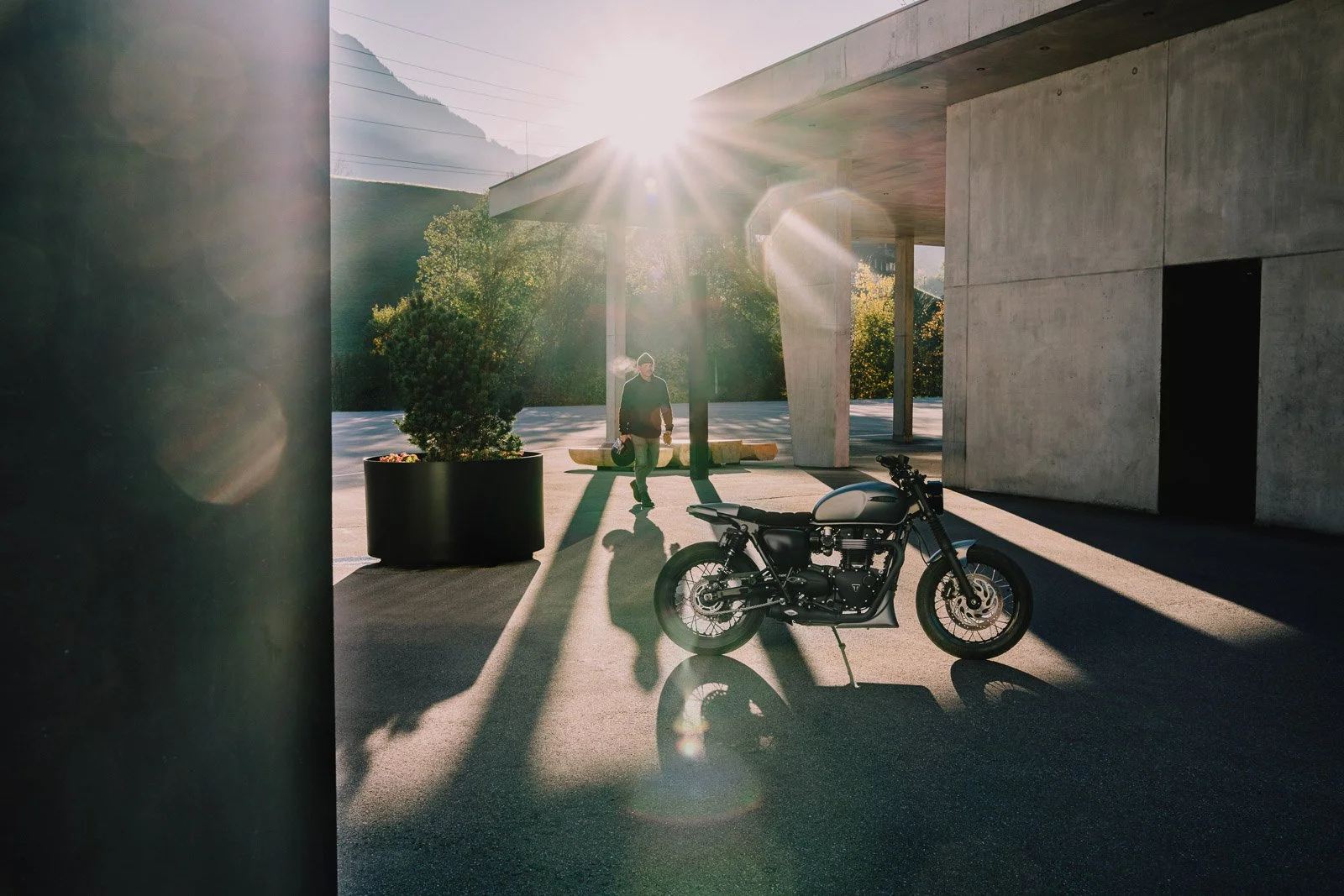 A man with a helmet in hand walking towards a parked motorcycle outside a modern concrete building, with the sun shining overhead creating lens flares and long shadows.