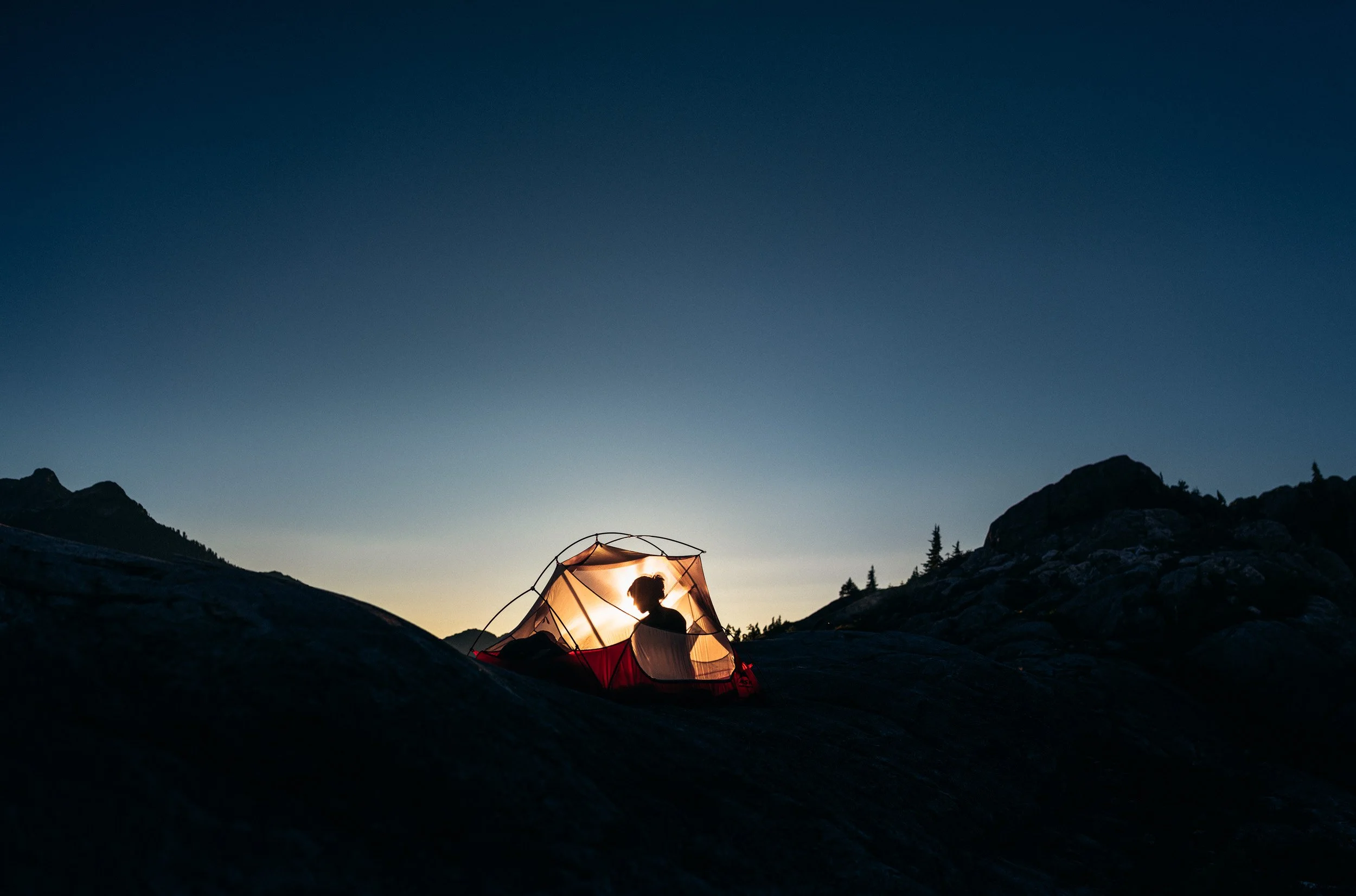 Person sitting inside a glowing tent on a rocky mountain landscape during dusk with a clear sky.