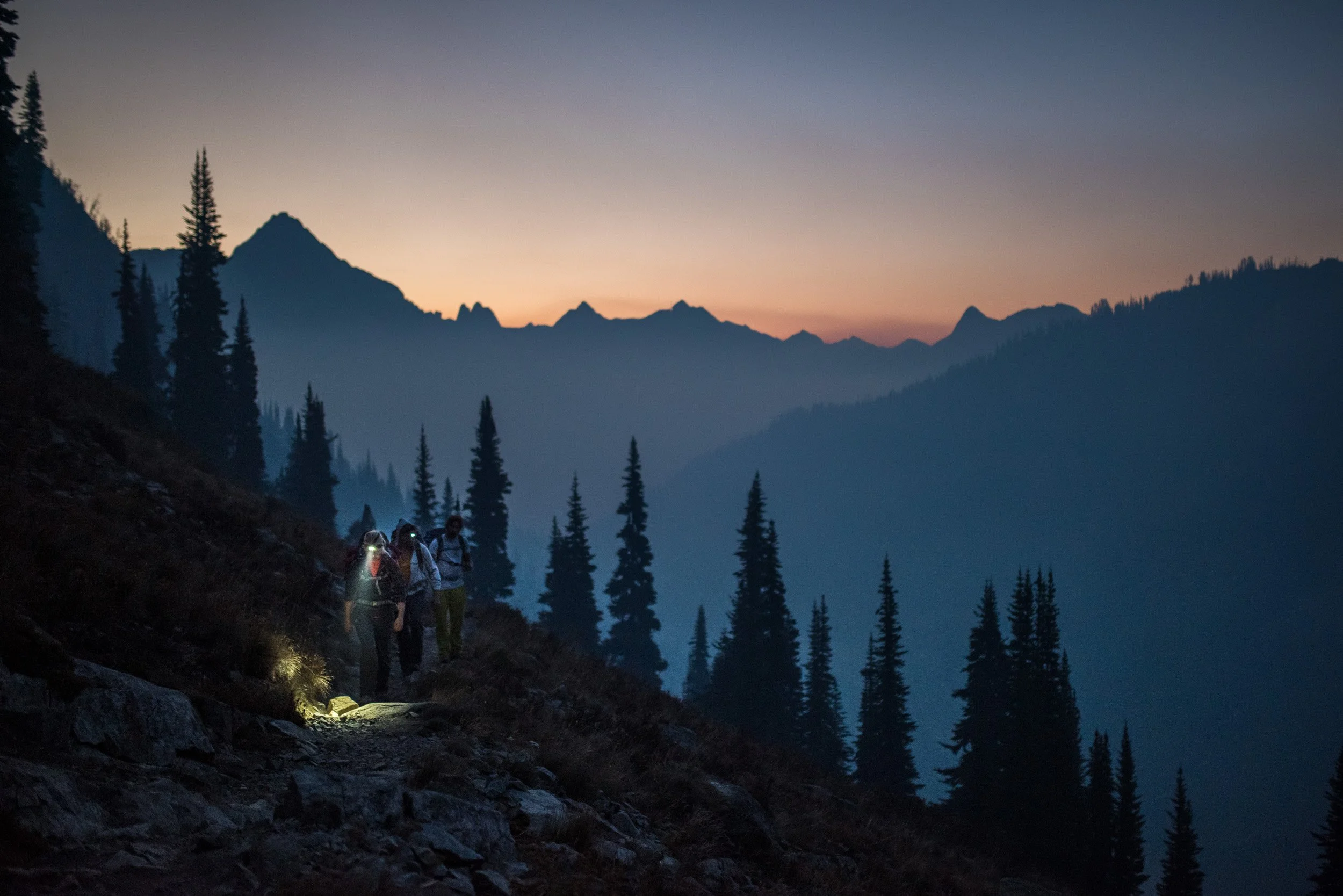 Hikers trekking on a mountain trail during dusk with pine trees and mountain silhouettes in the background.