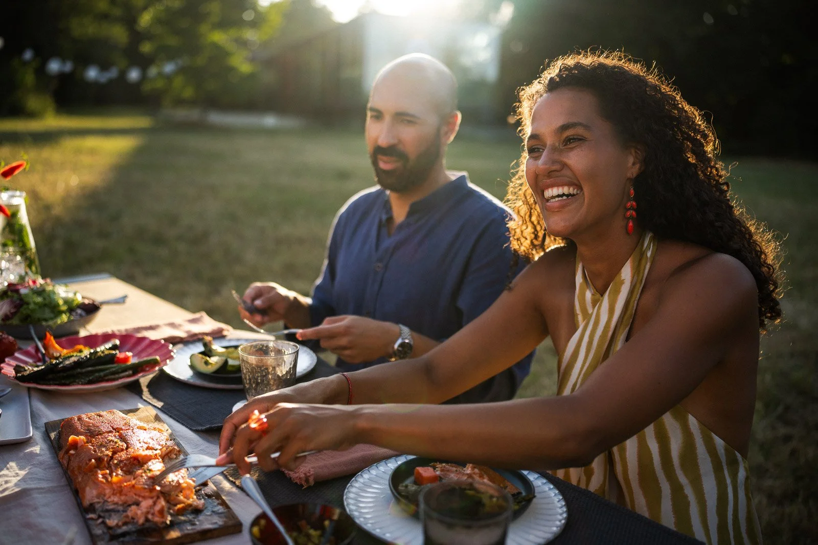 A woman with curly hair and earrings smiling while enjoying a meal at an outdoor dinner with a man in the background, with a table of food on a sunny evening.