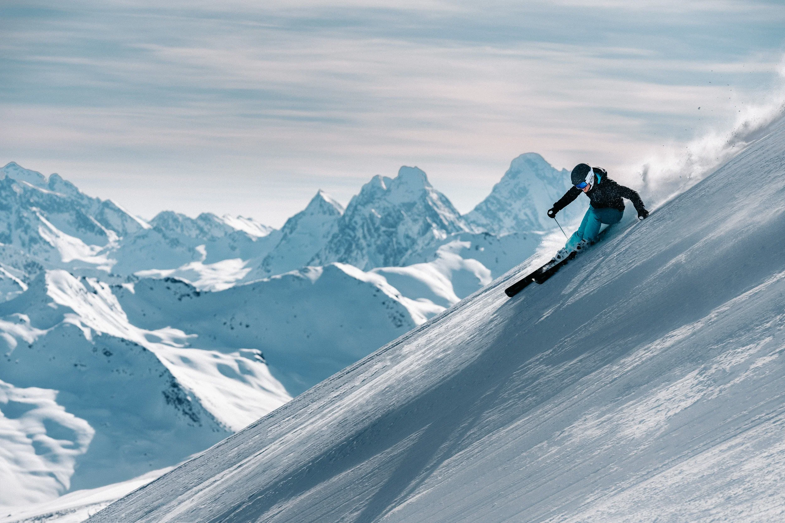 A person skiing downhill on a snowy mountain slope with a backdrop of snow-covered mountains under a cloudy sky.