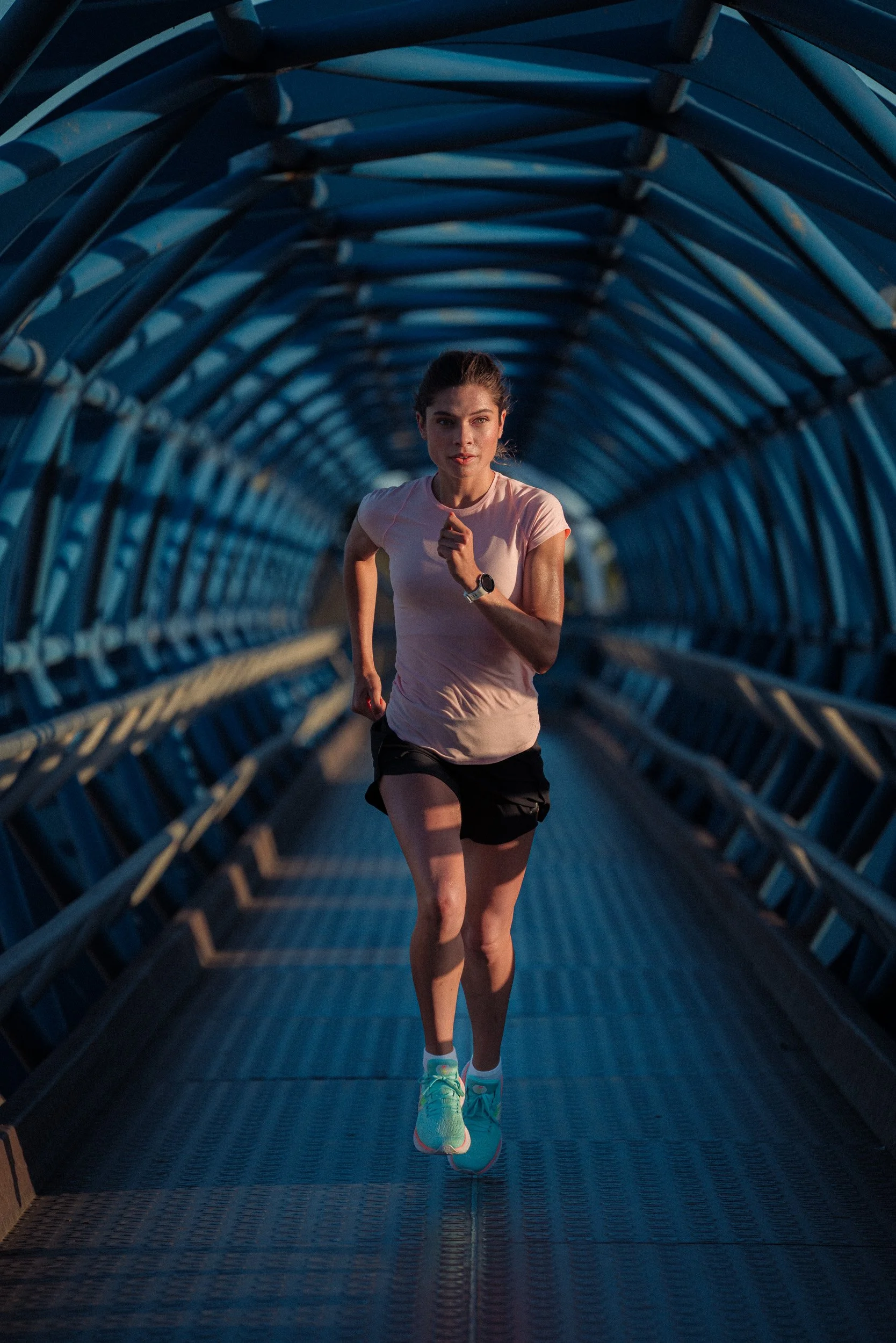 A young woman running through a tunnel on a bridge or walkway, wearing athletic clothes and a wristwatch, during what appears to be early morning or evening.