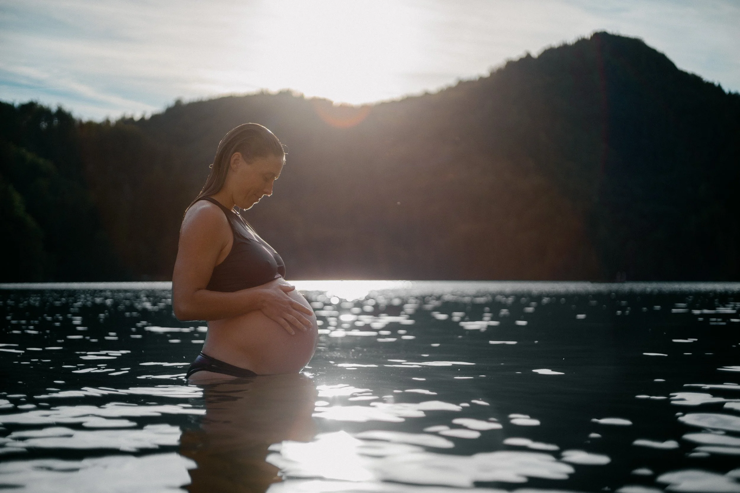 Pregnant woman standing in water during sunset, touching her belly