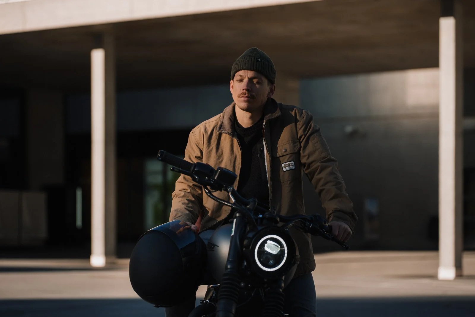 A man wearing a brown jacket and black beanie sitting on a motorcycle with a black helmet, outdoors during dusk or dawn.