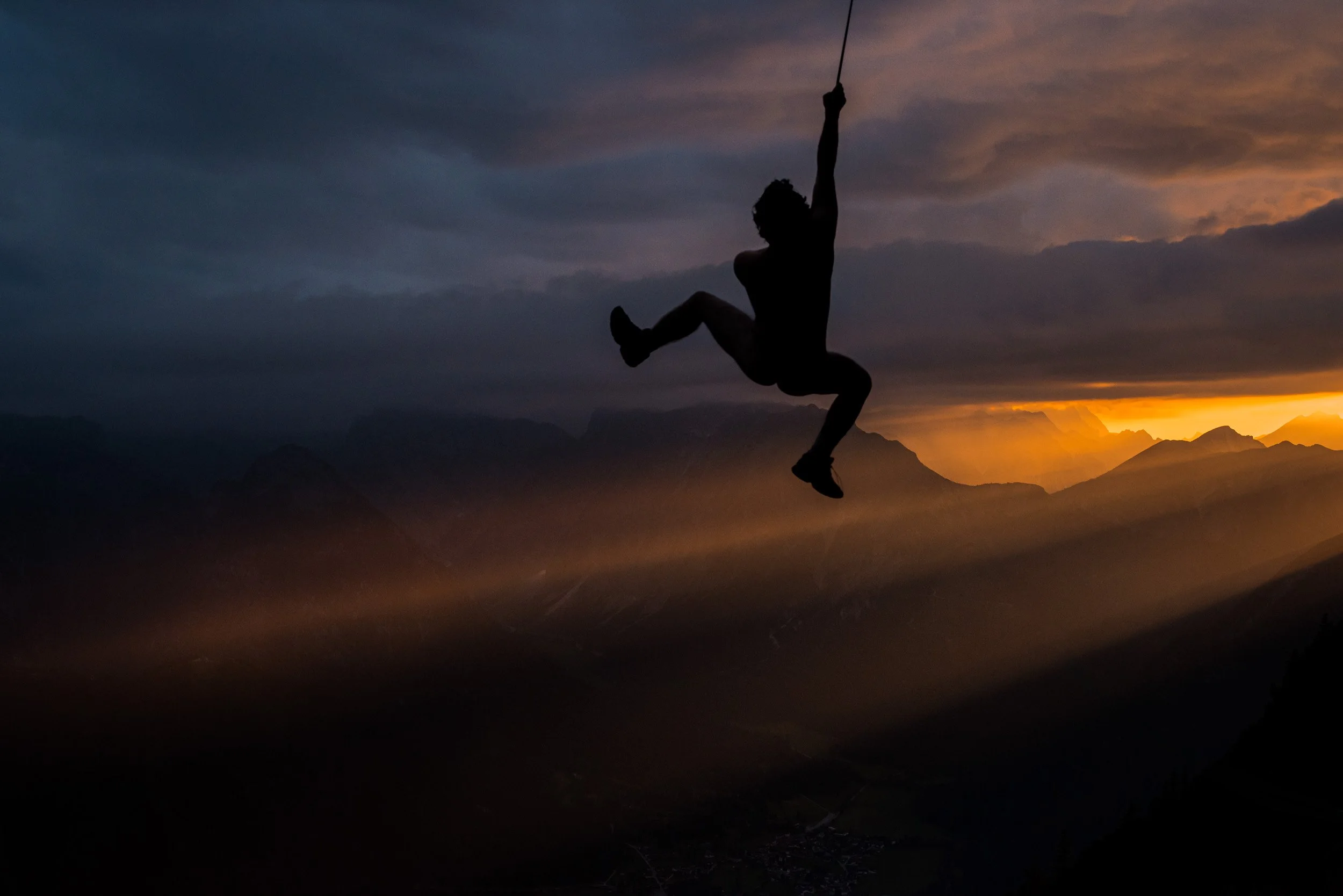 Silhouette of a person on a zip line against a sunset over mountain peaks with dark clouds in the sky.