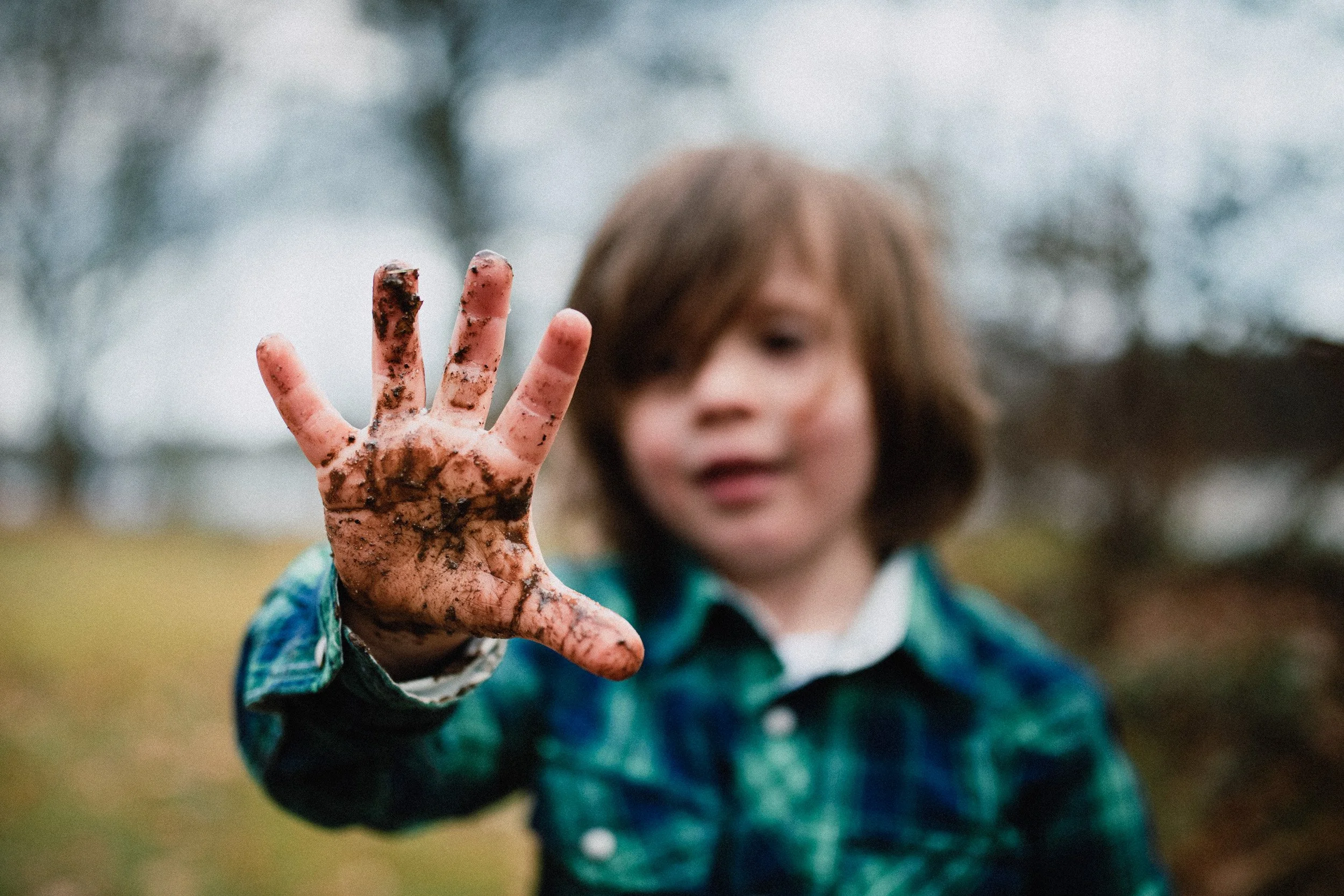 A young child with mud-covered hand extended toward the camera in an outdoor setting with trees in the background.
