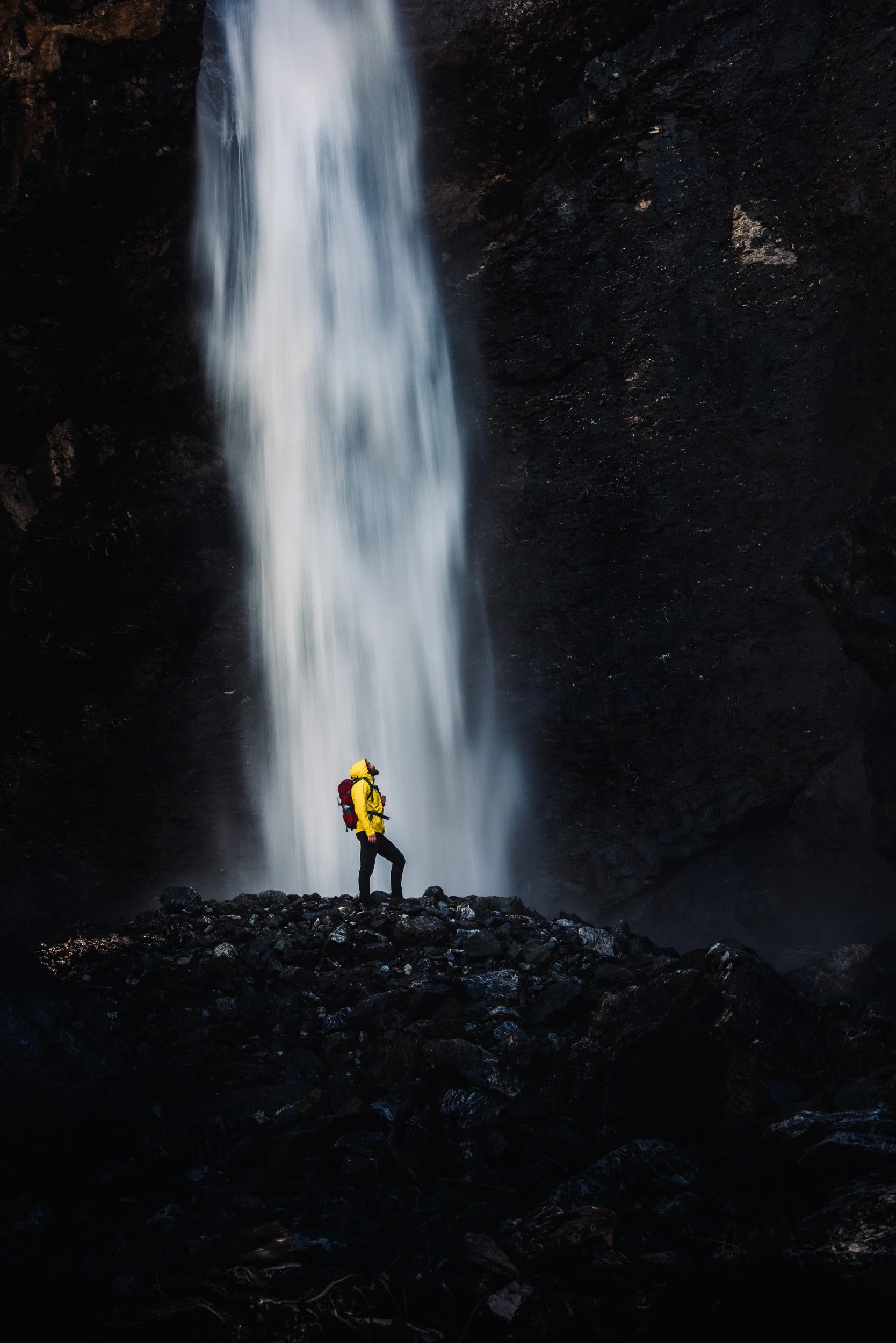 A person in a yellow jacket and backpack standing on rocks near a tall waterfall.