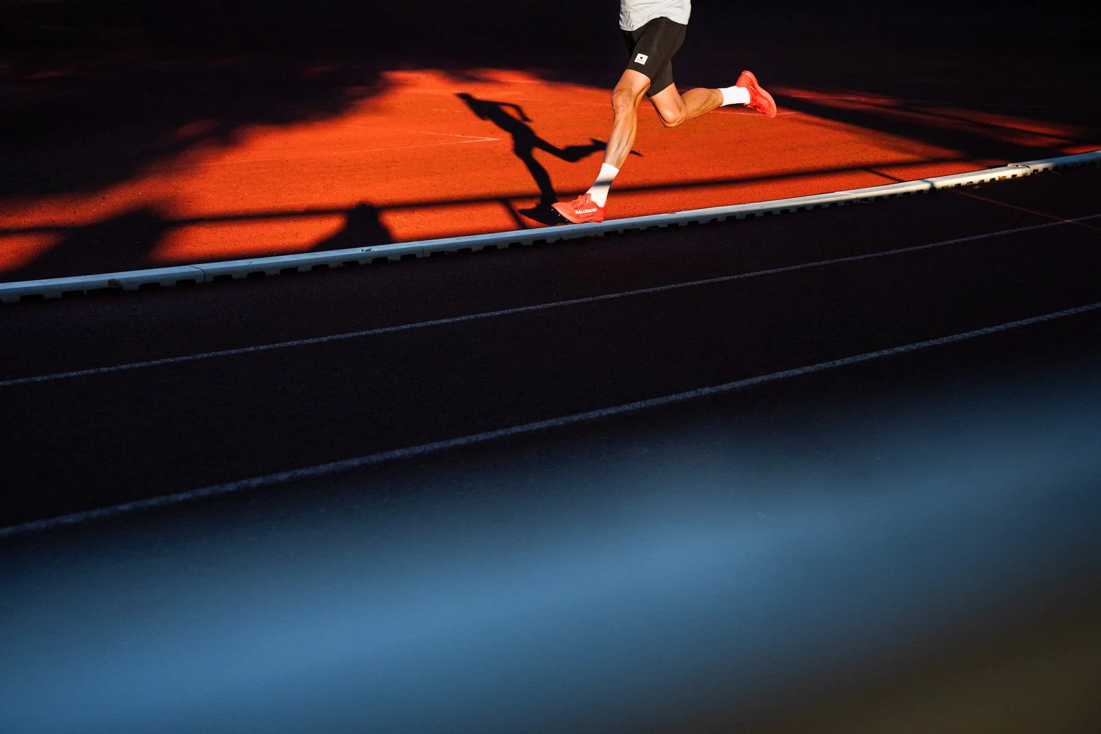 Runner in mid-stride on track, shadow cast on red surface, partial view, focused on lower body and legs