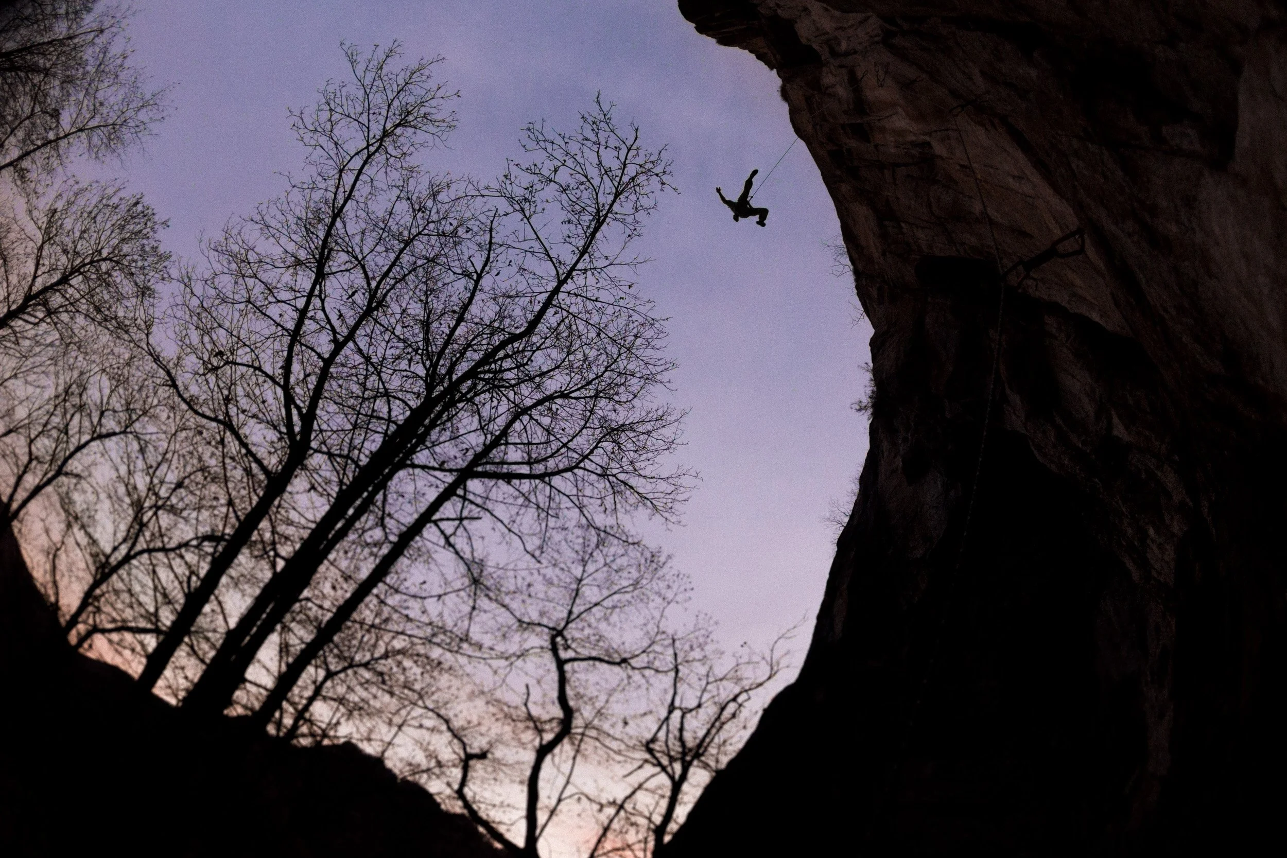 Person rock climbing on a cliff at sunset, silhouetted against a purple sky with leafless trees nearby.
