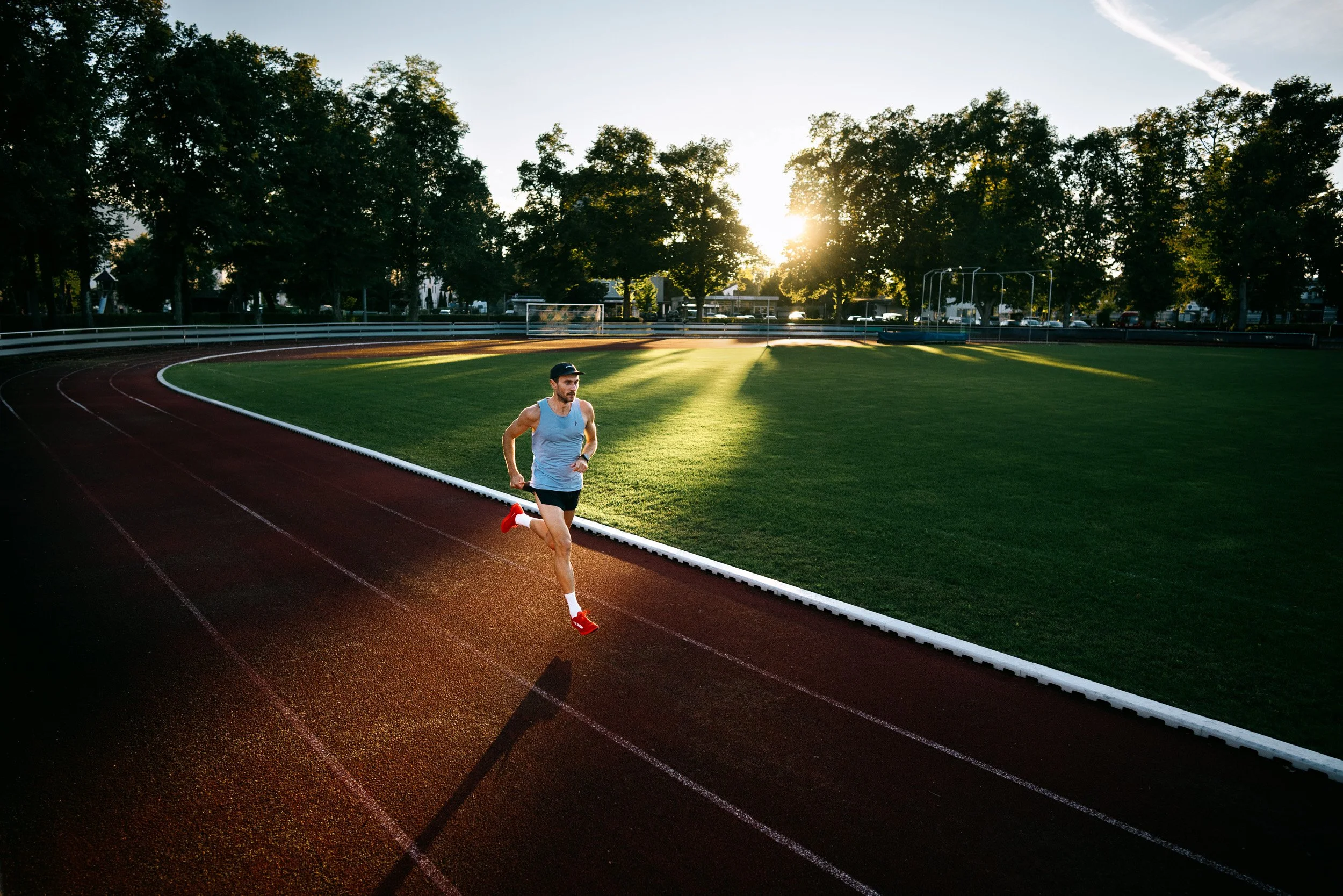 A man running on a red outdoor track at sunset with trees and a green field in the background.