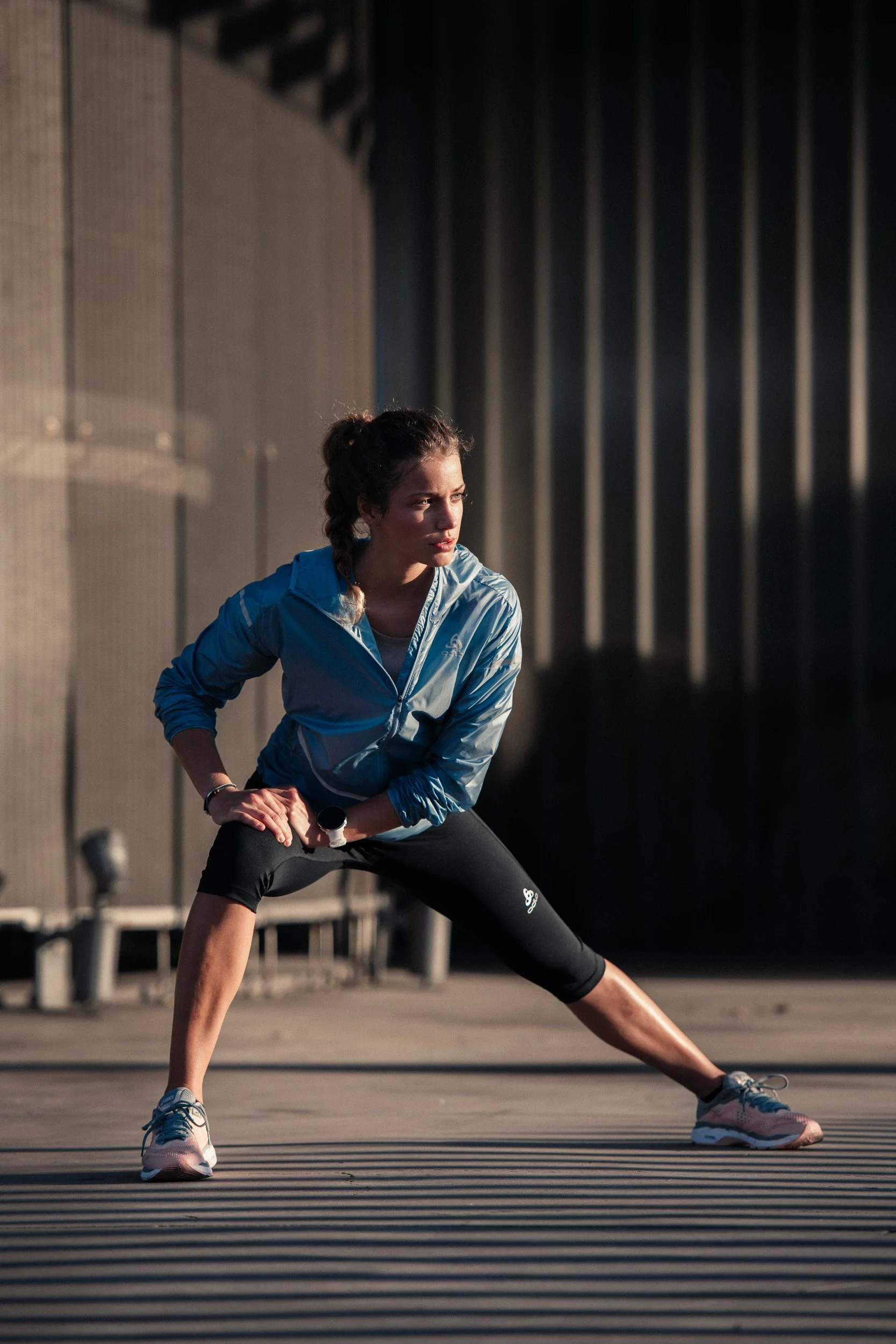 A woman in athletic wear stretching outdoors in a pose with legs apart, wearing a blue jacket, black leggings, and pink running shoes, with a background of a building and shadow lines.