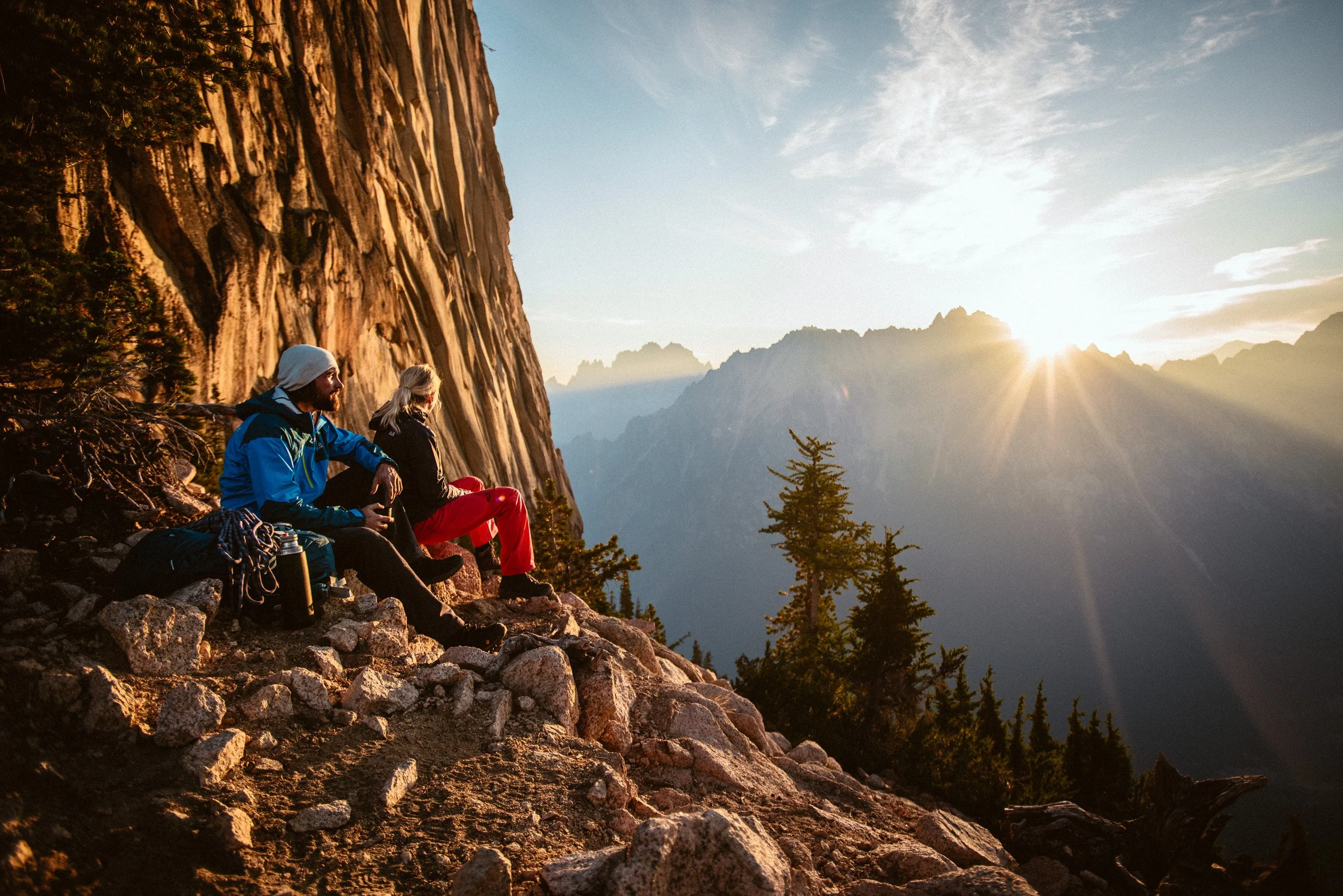 Two hikers sitting on rocky terrain with mountains and trees in the background during sunset.