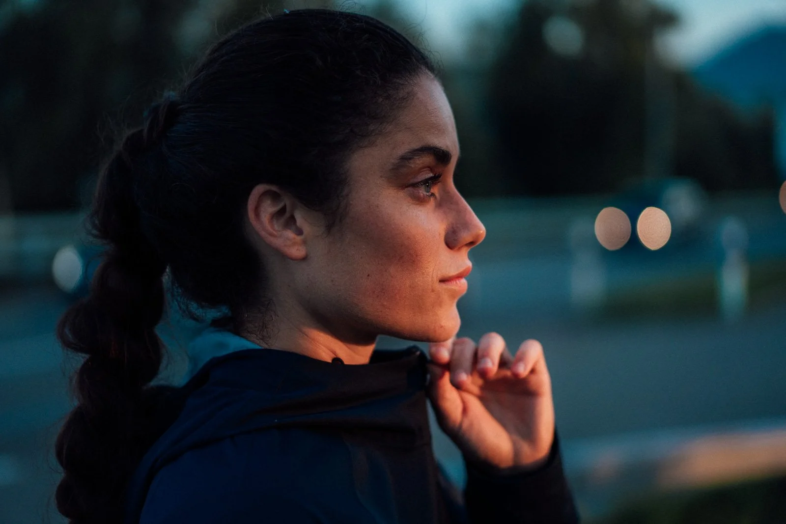 Close-up of a woman with dark hair in a ponytail, wearing a dark sports jacket, standing outdoors during sunset, looking to the right with a slight smile.