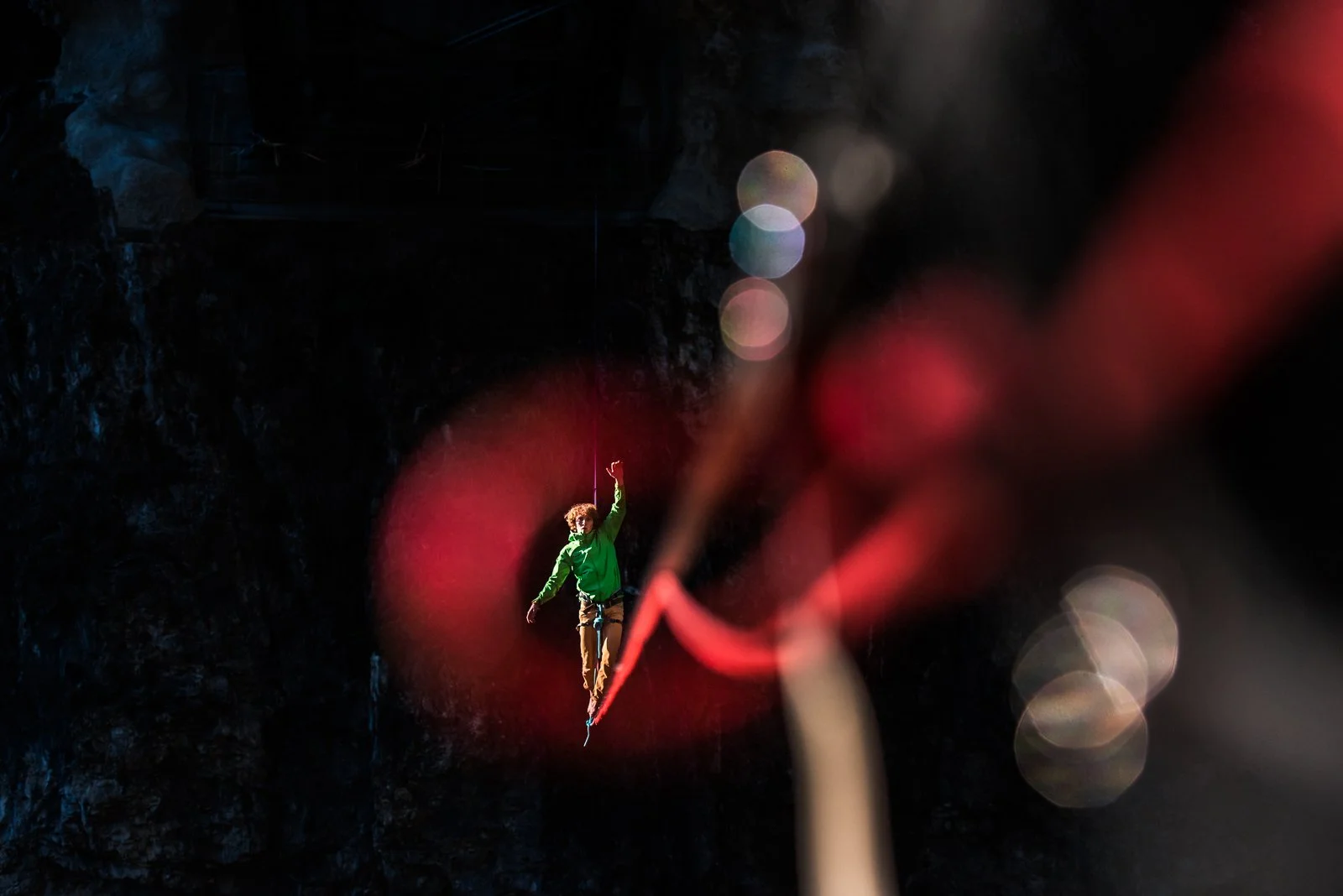 A person in a green jacket walking on a tightrope with a harness. The background is dark and blurry with bokeh lights, and the foreground has out-of-focus red ropes or cables.