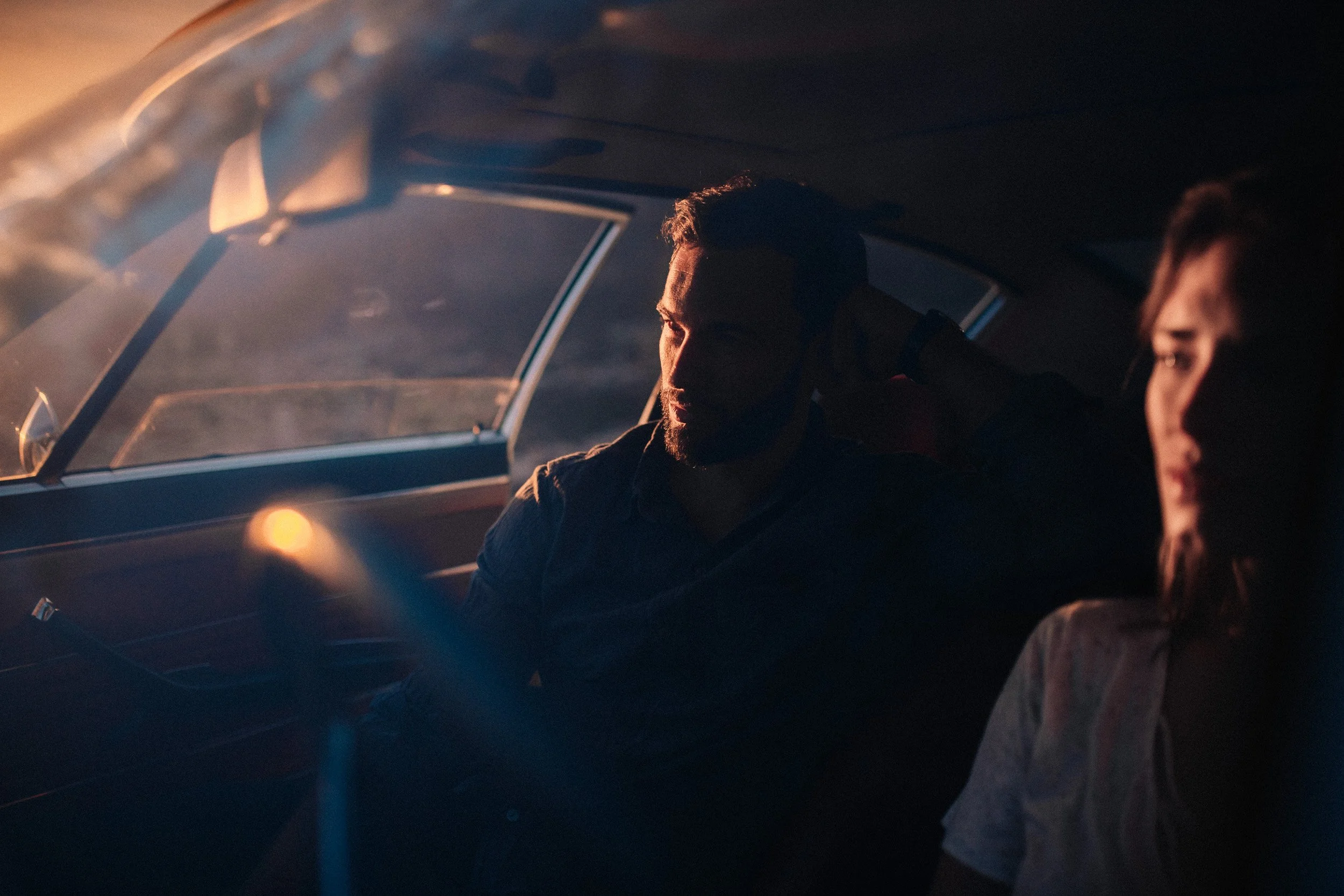A man and woman sitting inside a car at sunset, with the man looking outside the window and the woman looking downward, both appearing contemplative.