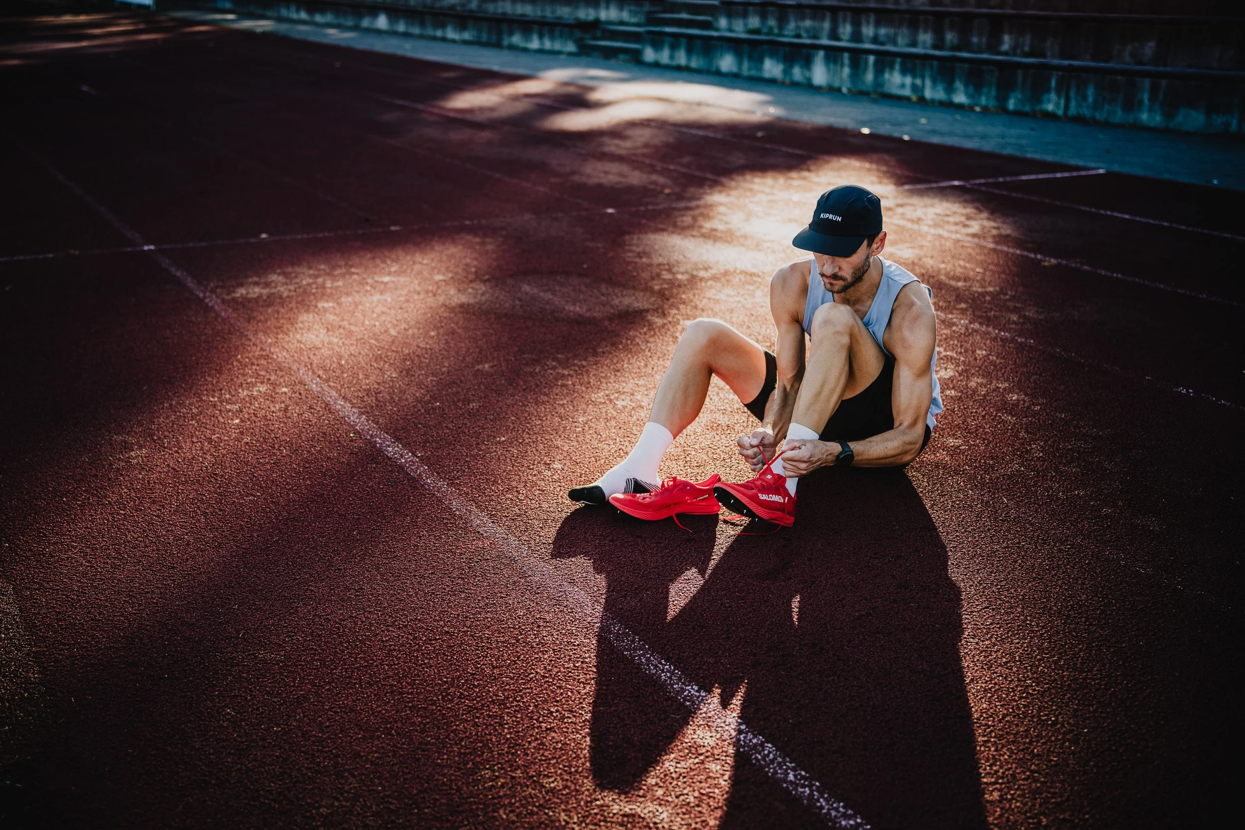 A male runner wearing a black cap, sleeveless gray shirt, black shorts, and white socks with black stripes, sitting on a red running track tying his red running shoes.
