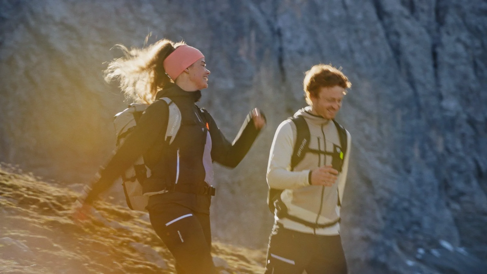 Two people, a woman with curly hair and a man with short hair, trail running on a mountain trail with a rocky background, both wearing outdoor gear and backpacks, smiling and enjoying the activity.