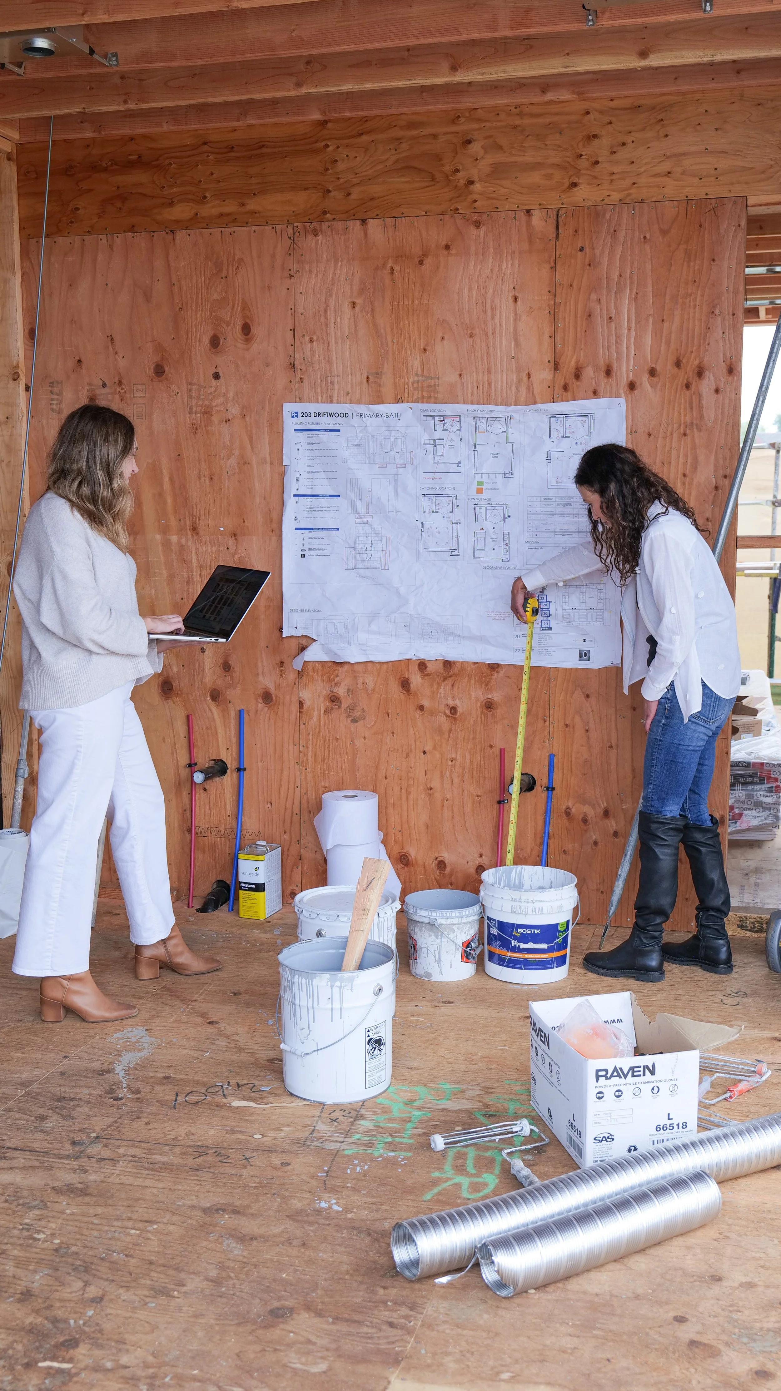 Two women working on a construction project inside a building with wooden framing. One woman is measuring a large blueprint on the wall, while the other holds a laptop. Construction materials, buckets, and tools are scattered on the wooden floor.