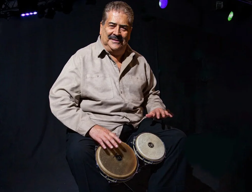 Johnny "Dandy" Rodríguez later in life, smiling, with a pair of bongos on his knees