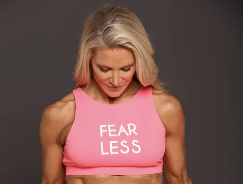 A woman with blonde hair wearing a pink sports bra with the words 'FEAR LESS' printed on it.