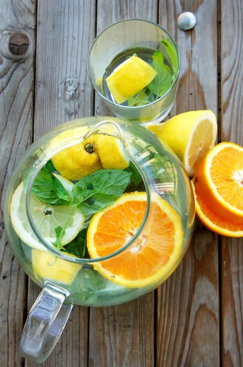 A glass pitcher with lemon, orange slices, and fresh green basil leaves on a wooden surface, with a glass of water containing lemon and basil nearby.