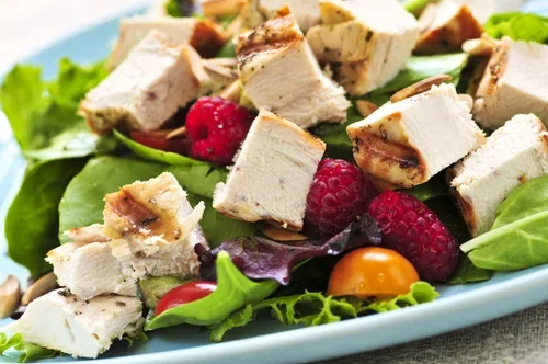 Salad with lettuce, raspberries, cherry tomatoes, and grilled chicken pieces in a blue bowl.