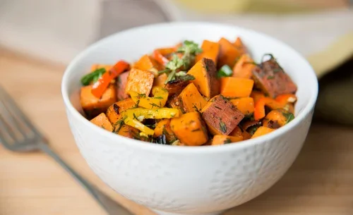 A white bowl filled with roasted sweet potatoes, carrots, and herbs on a wooden surface.