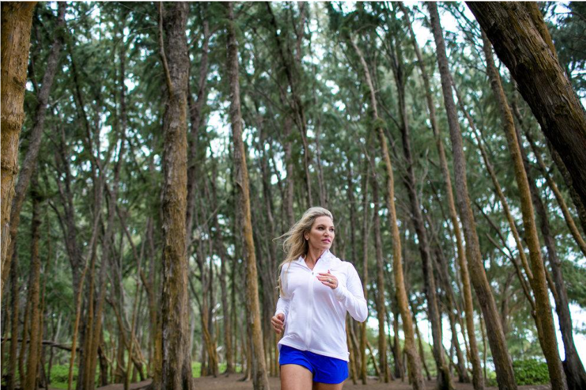 A woman jogging in a forest with tall trees and green foliage.