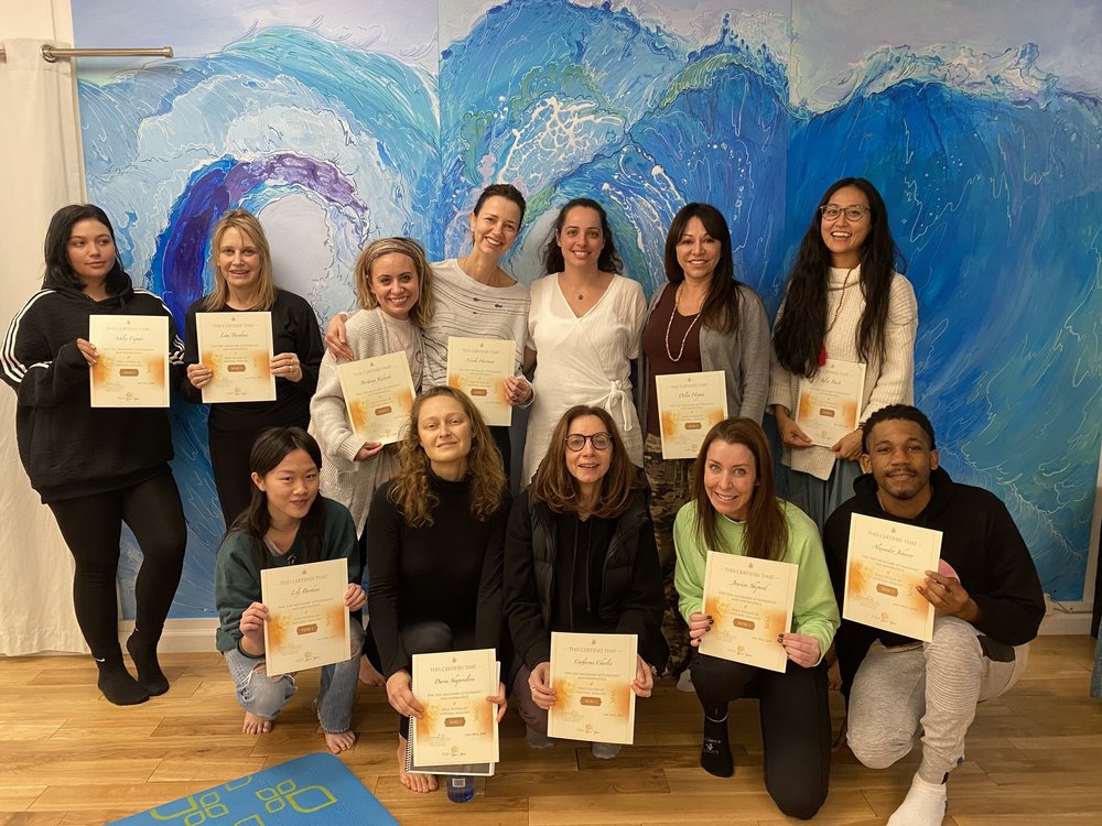 A training group holding their certificates in front of the painted blue wave that covers one wall of the Atma studio