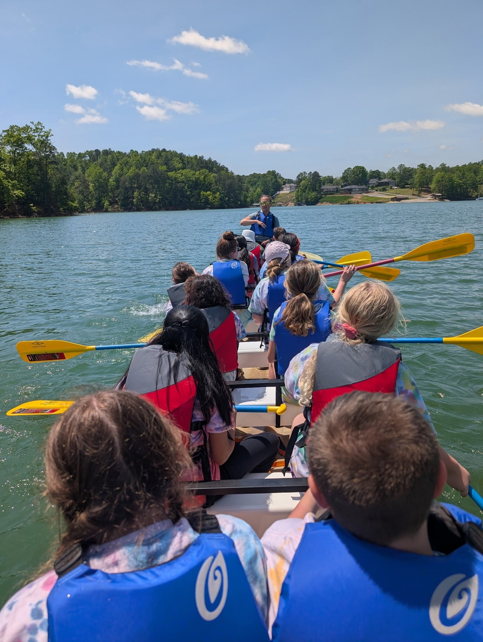 Students wearing life jackets hold yellow paddles in a large canoe, viewed from behind, with a wooded shoreline ahead