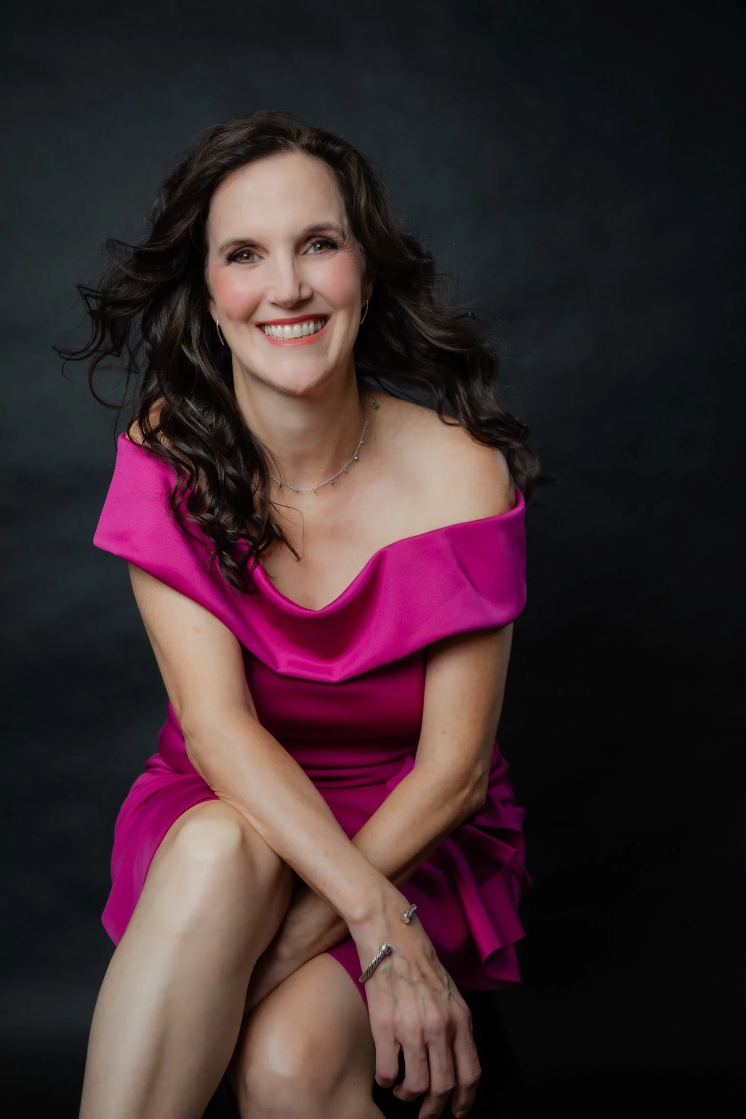Portrait of a woman with dark hair wearing a pink of-the-shoulder dress on a black background.