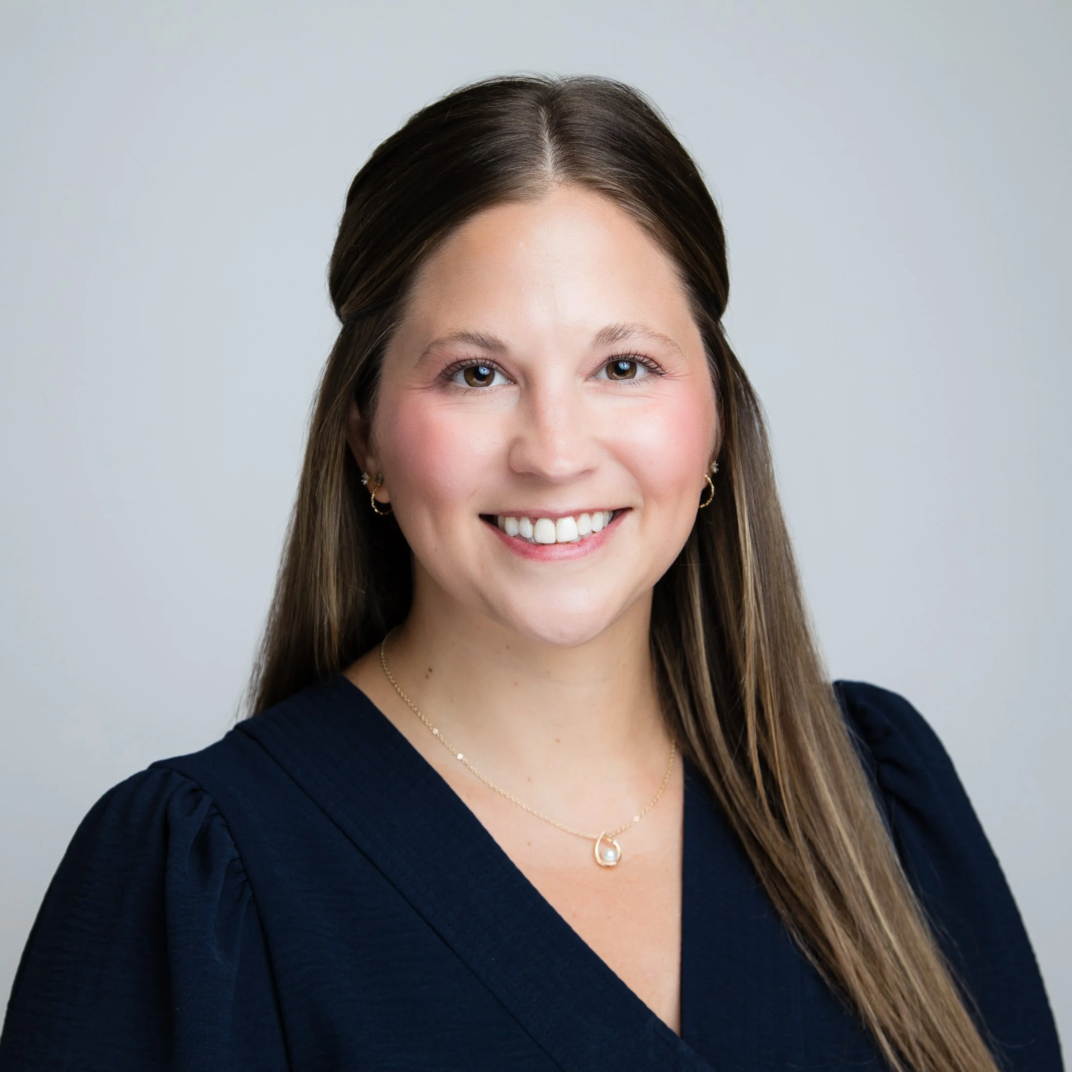 Headshot of a woman with straight brown hair and green eyes, wearing a navy blouse on a light gray background. Photographed in studio by Amanda Faucett Photography.